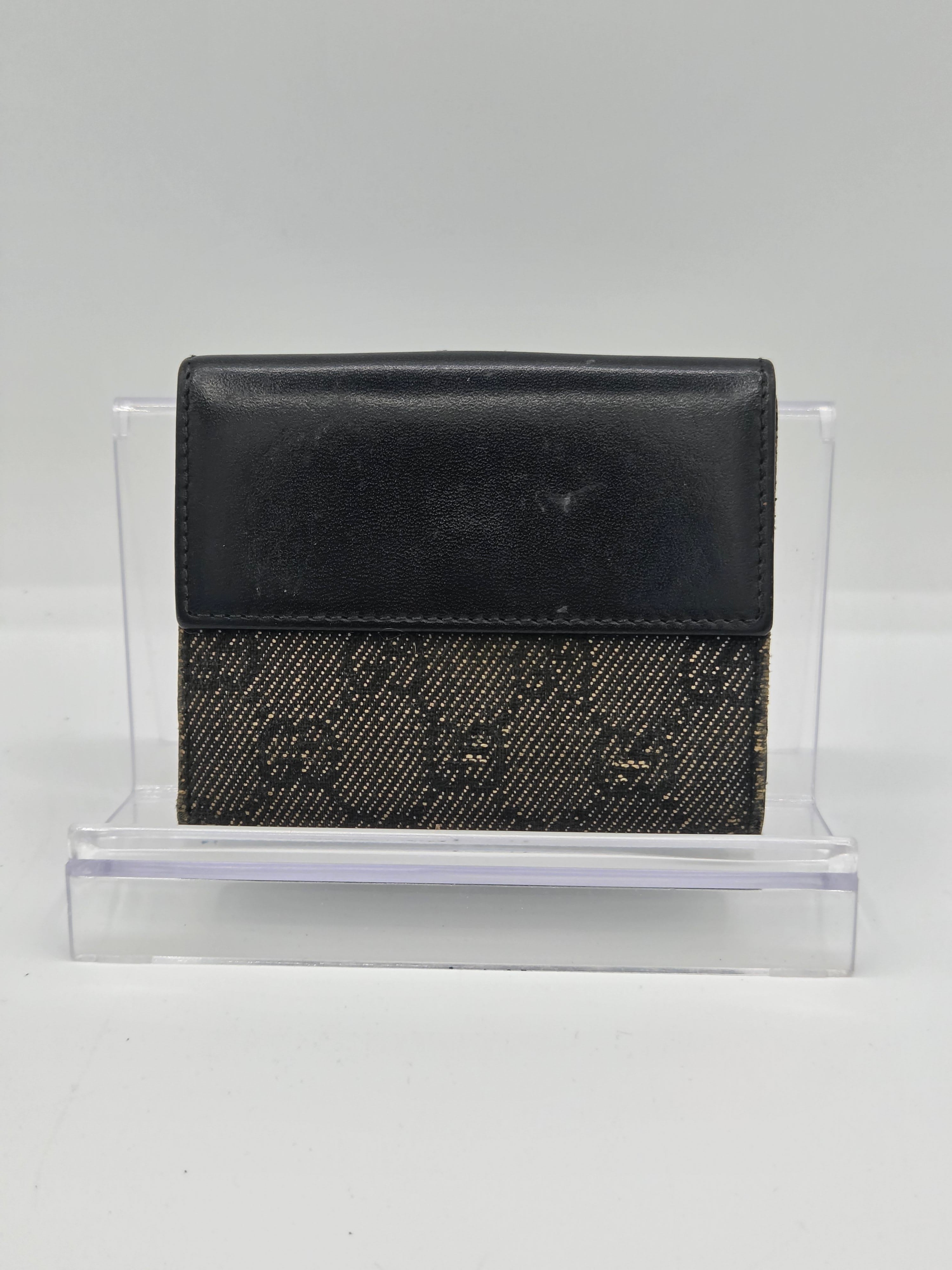 A black leather and monogram canvas bi-fold wallet displayed in a clear acrylic stand against a plain white background.
