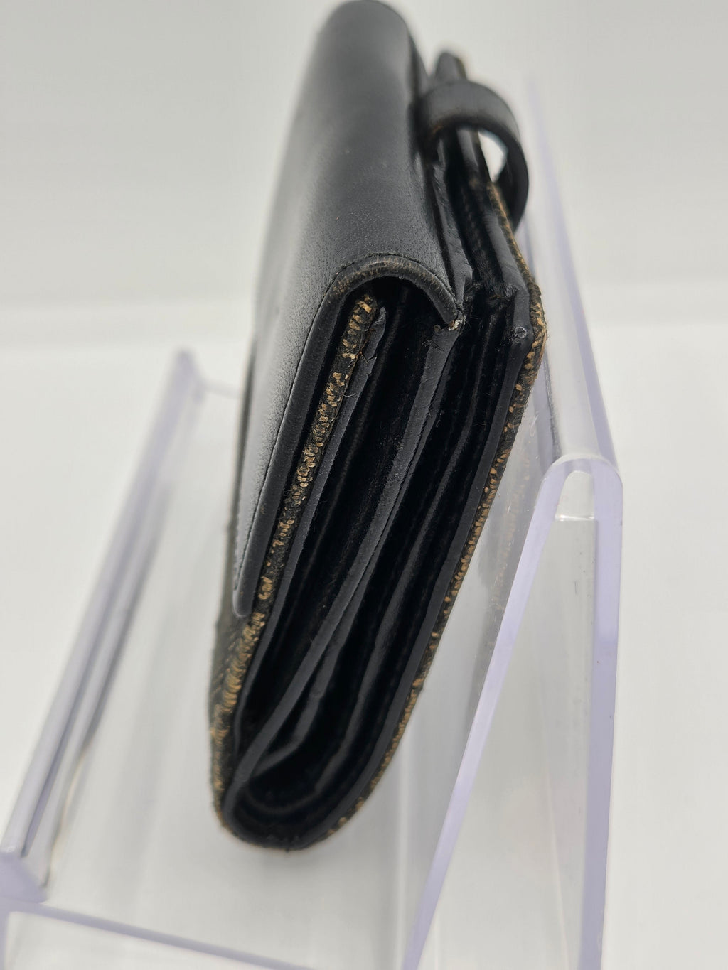 A close-up of a black leather wallet or card holder with gold glitter trim, shown resting on a clear acrylic display stand against a white background.