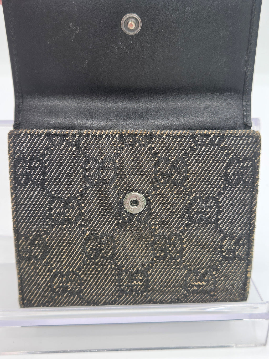 A close-up of a black and dark gray woven monogram wallet or card holder, shown open with a black leather interior on a clear display stand.