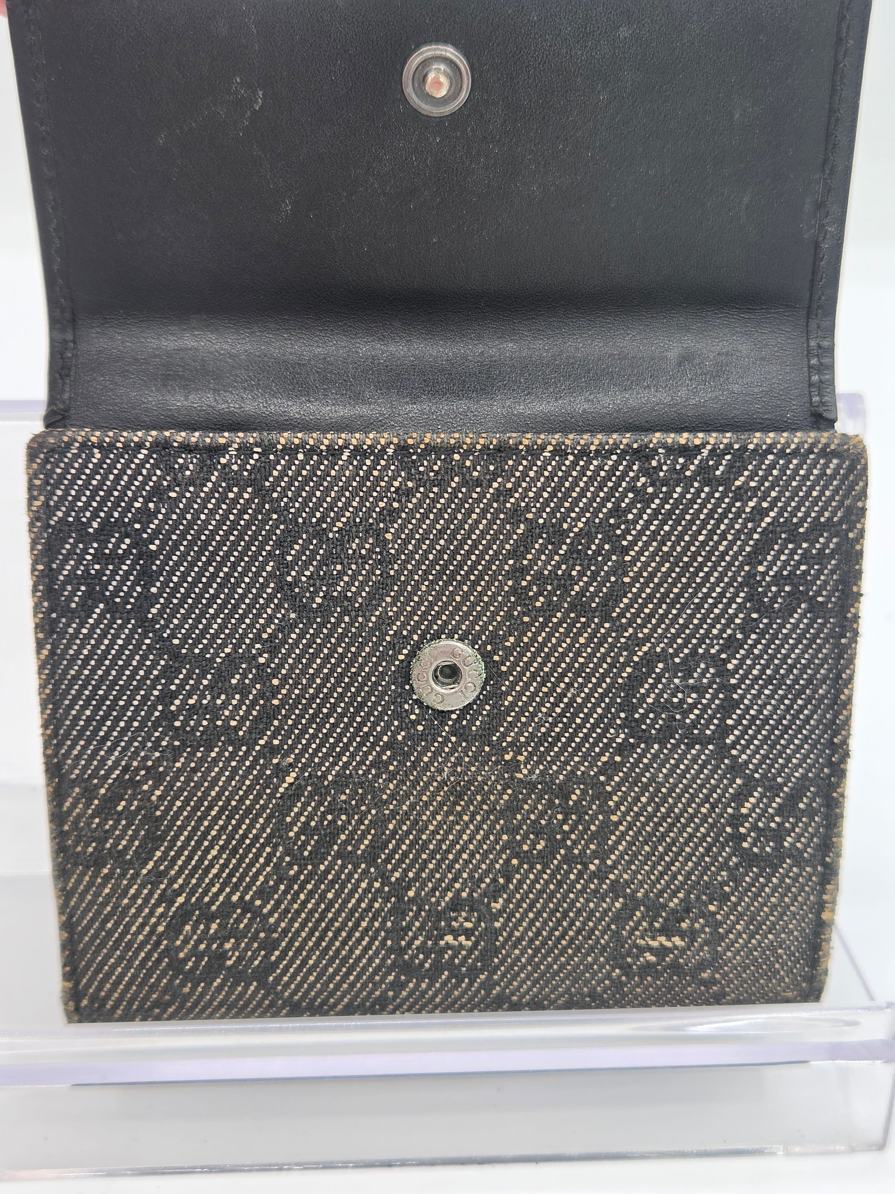 A close-up of a black and dark gray woven monogram wallet or card holder, shown open with a black leather interior on a clear display stand.