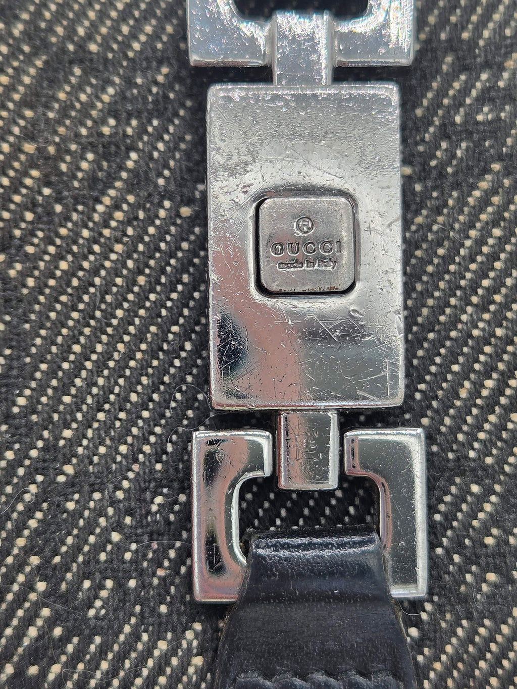 Close-up of a silver-toned metal clasp on a textured black and beige woven fabric, with a black strap visible on the right.