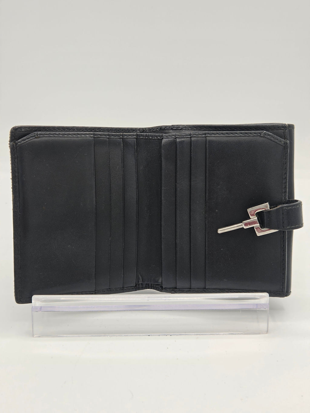 A black leather wallet with a silver-toned metal clasp closure, shown open next to a clear plastic protective sleeve on a plain white background.