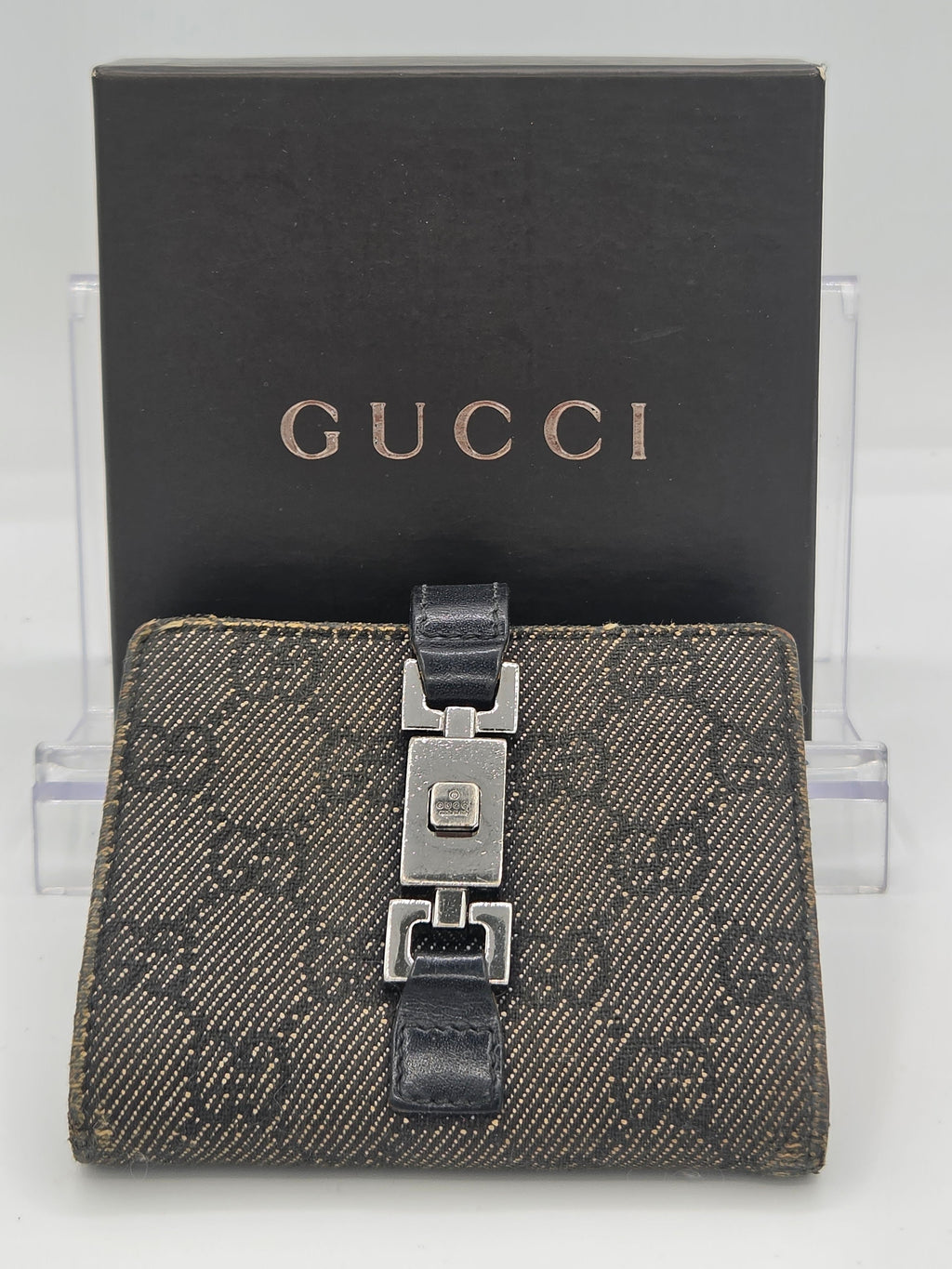 A Gucci monogram wallet with a dark brown and black GG Supreme canvas body, a black leather strap, and a silver-tone metal clasp, displayed in front of its black Gucci box on a clear acrylic stand.