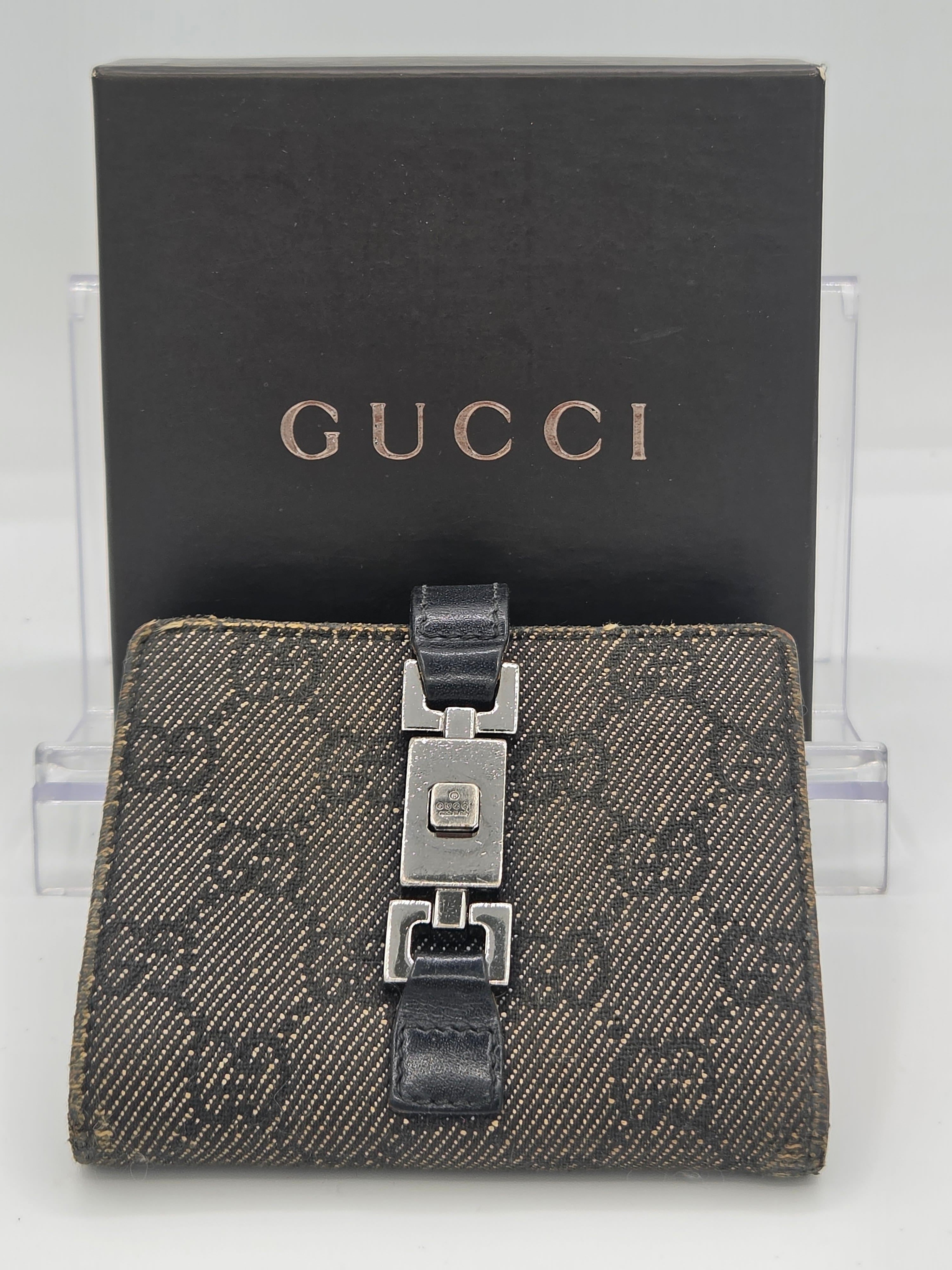 A Gucci monogram wallet with a dark brown and black GG Supreme canvas body, a black leather strap, and a silver-tone metal clasp, displayed in front of its black Gucci box on a clear acrylic stand.