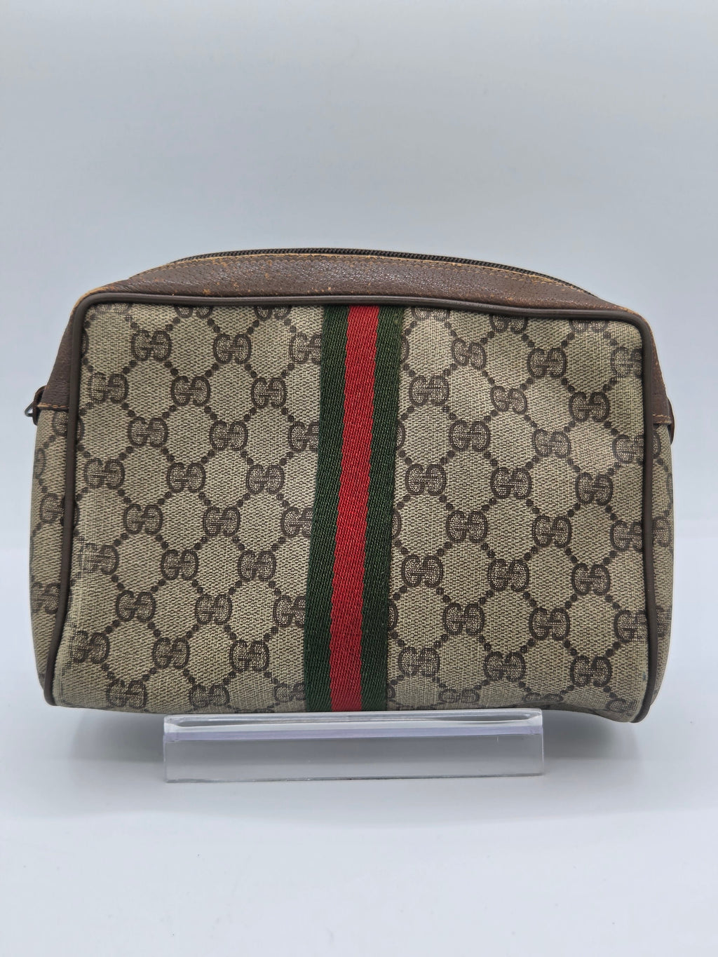 A Gucci monogram canvas pouch with a brown leather trim and a signature green and red web stripe, shown against a plain light gray background.