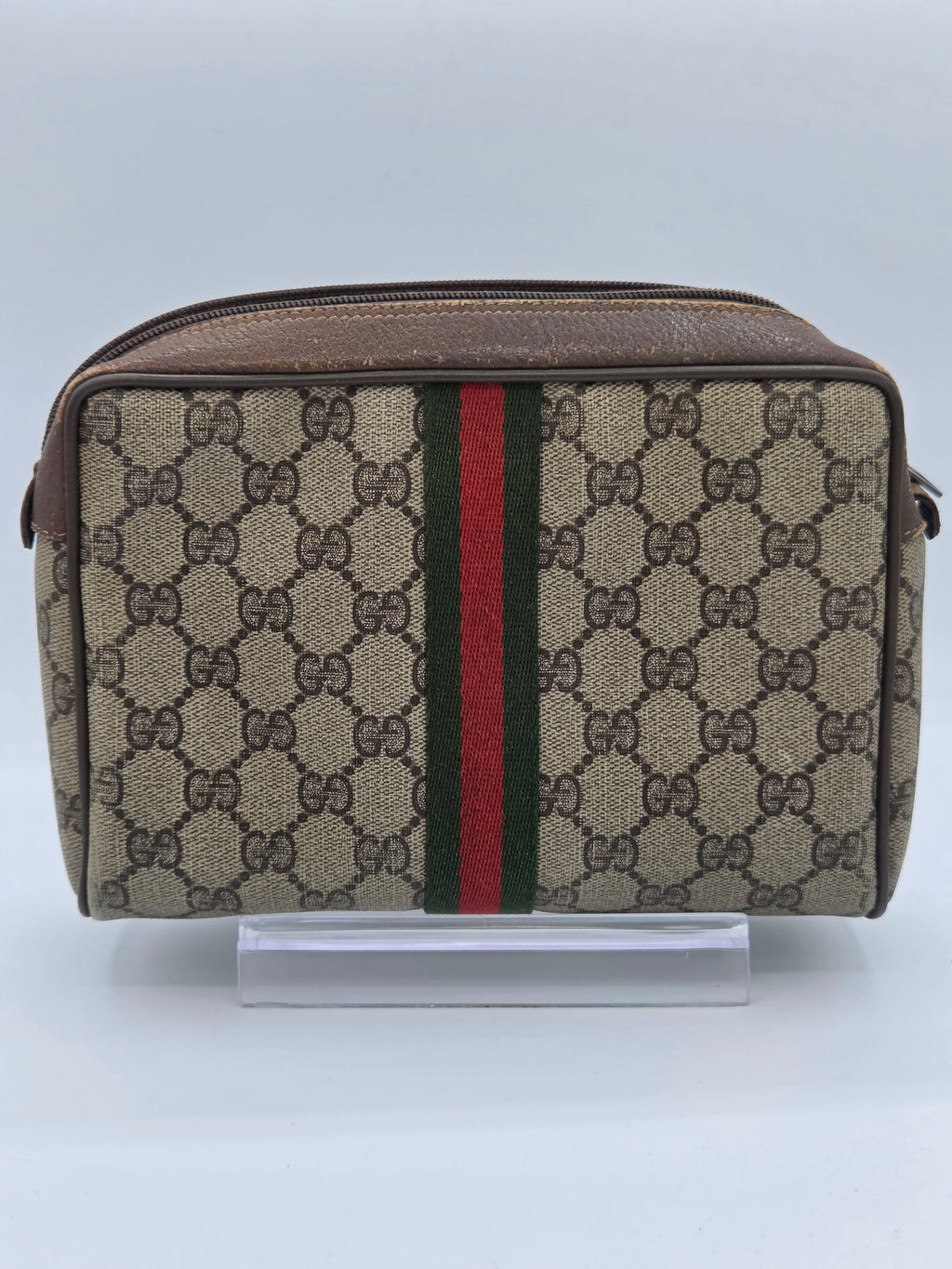 A Gucci monogram canvas pouch with a brown leather trim and the signature green and red web stripe, shown against a plain light background.