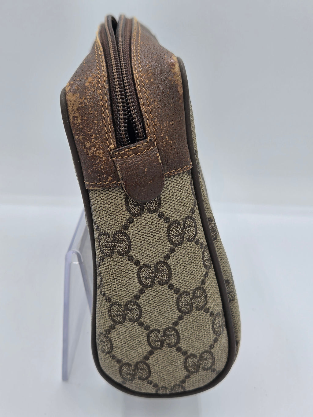 A Gucci zippered pouch in the brand's signature GG monogram canvas and brown leather, displayed on a clear stand against a white background.