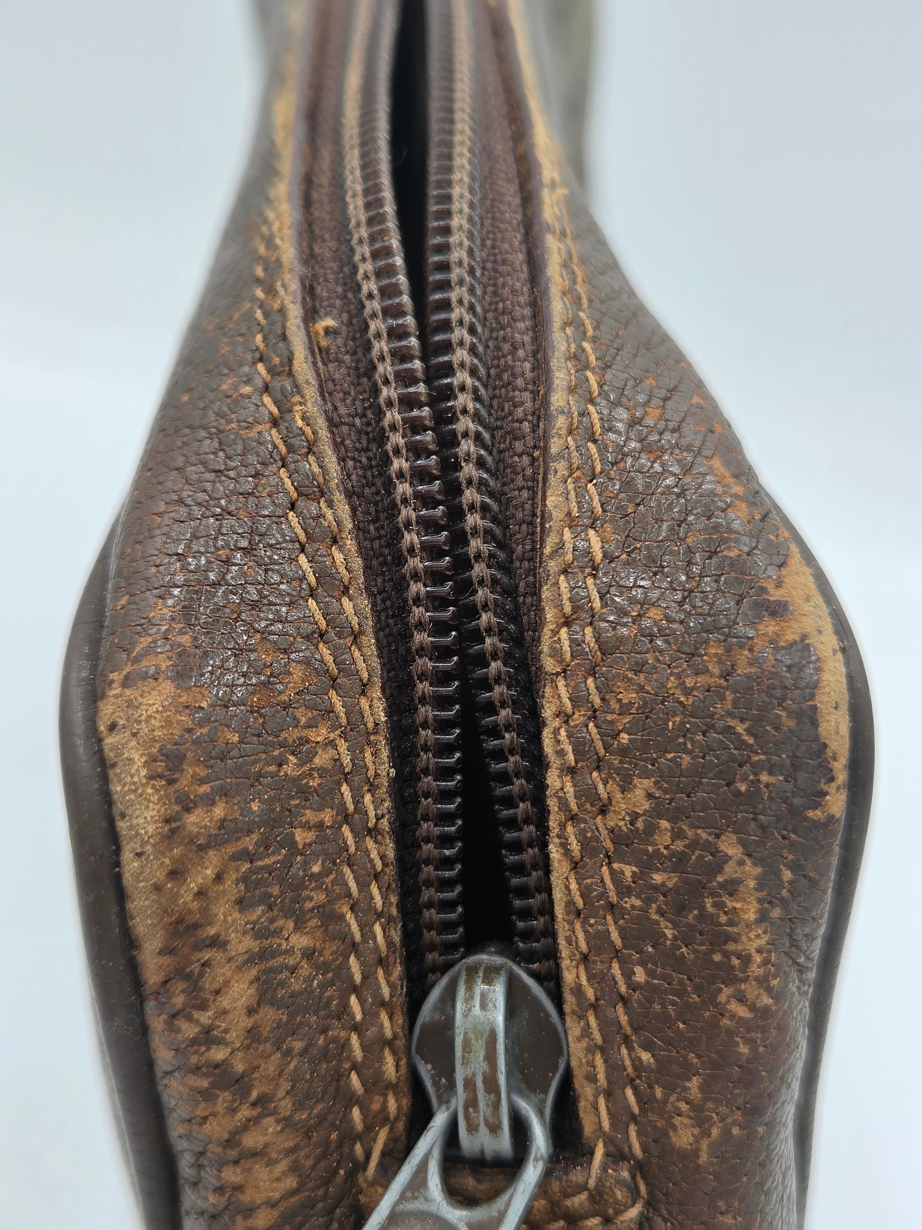 A close-up of a brown textured leather zippered pouch, showing the worn monogram pattern, stitching, and a silver metal zipper pull against a white background.
