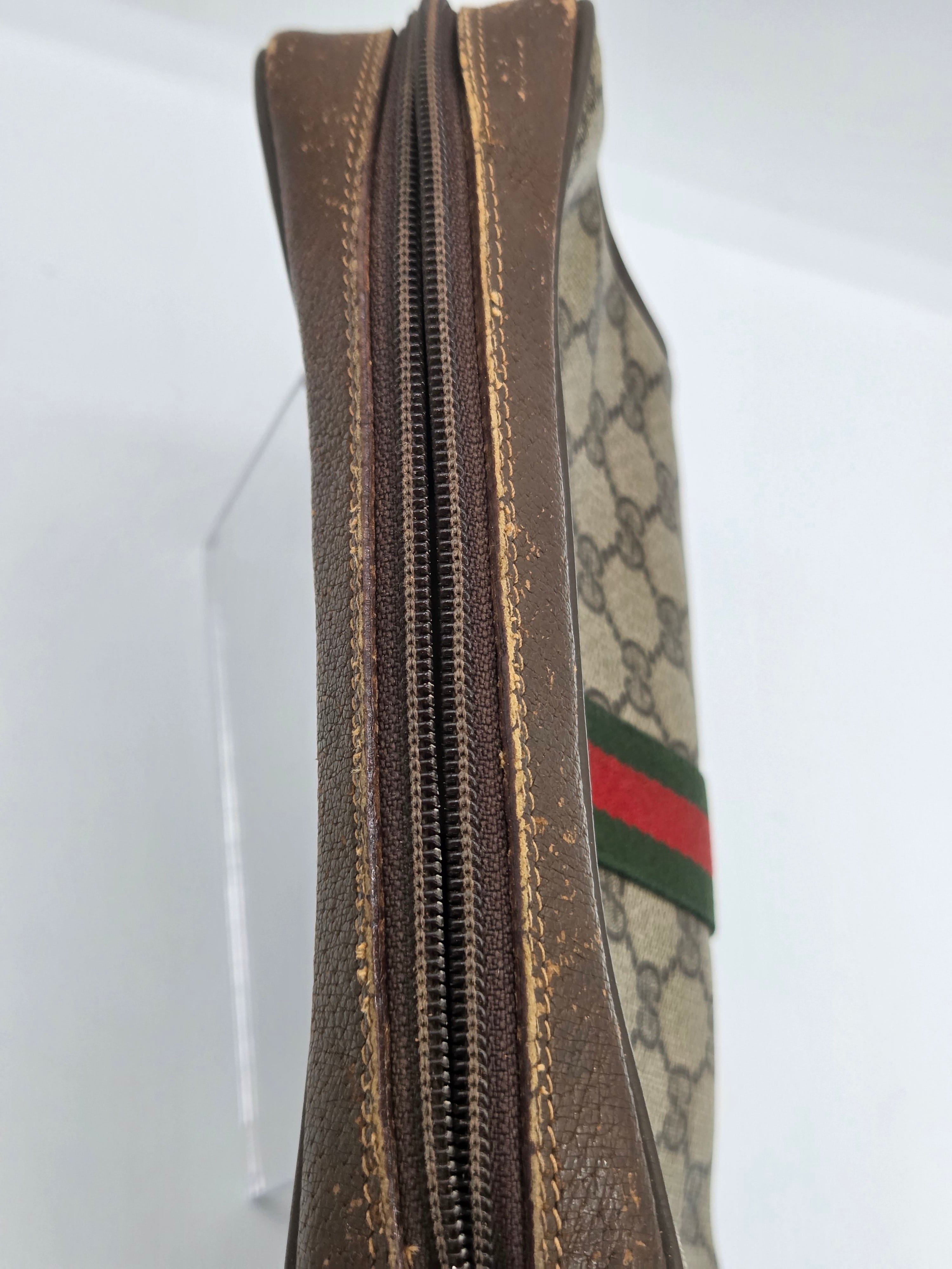 A close-up side view of a Gucci zippered pouch or wallet, showing the brown monogram canvas, distressed brown leather trim, and the signature green and red web stripe.