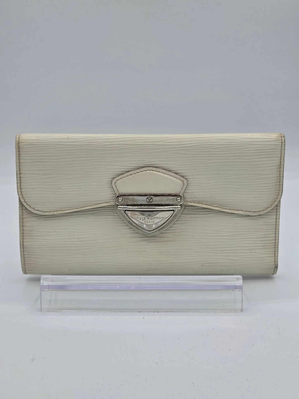 A light beige textured leather clutch bag with a silver-tone metal clasp, displayed next to a clear plastic display block on a light gray background.