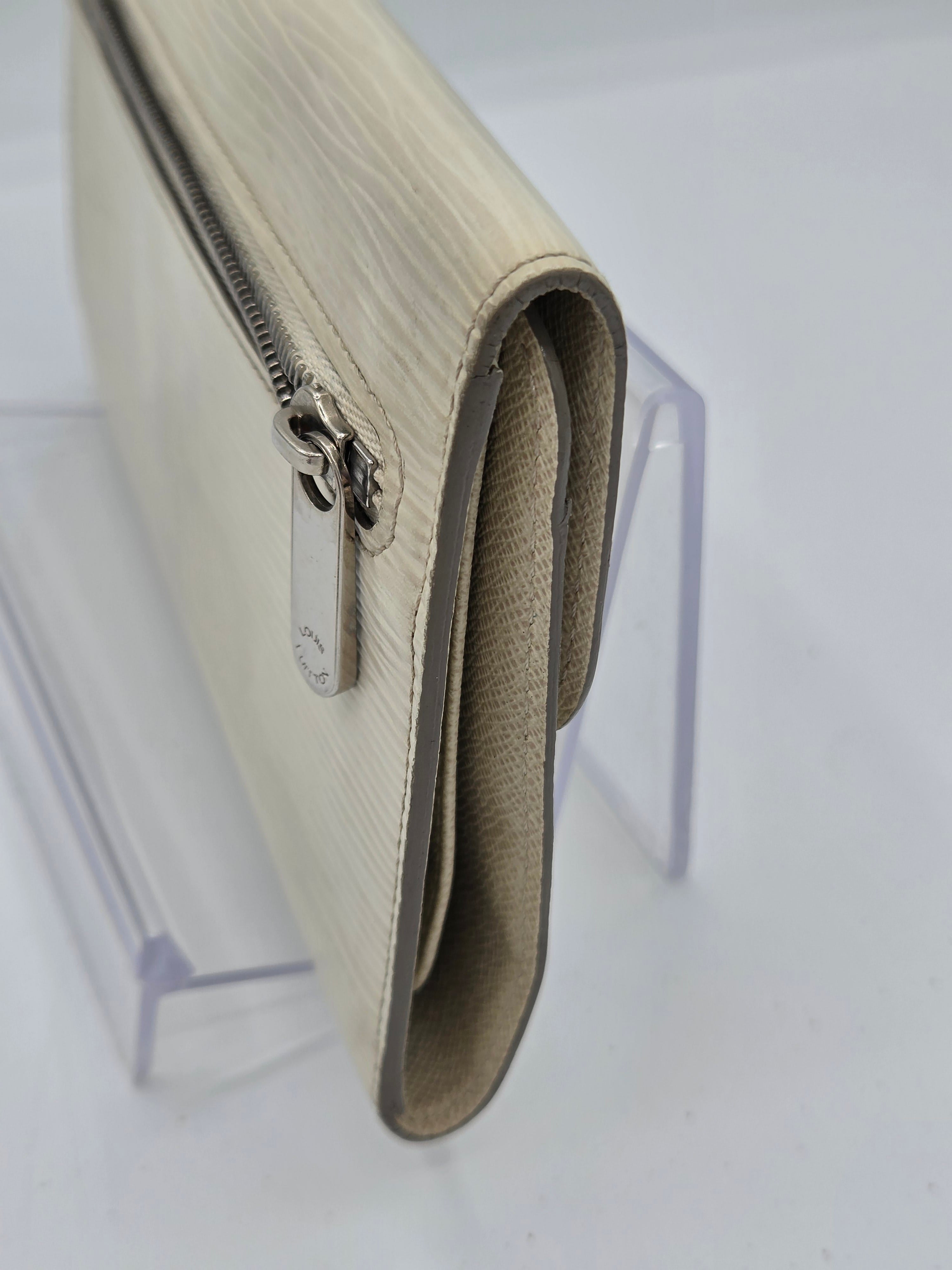 A close-up of a cream-colored textured leather wallet or clutch with a silver zipper and a small metal logo tab, resting on a clear acrylic display stand.