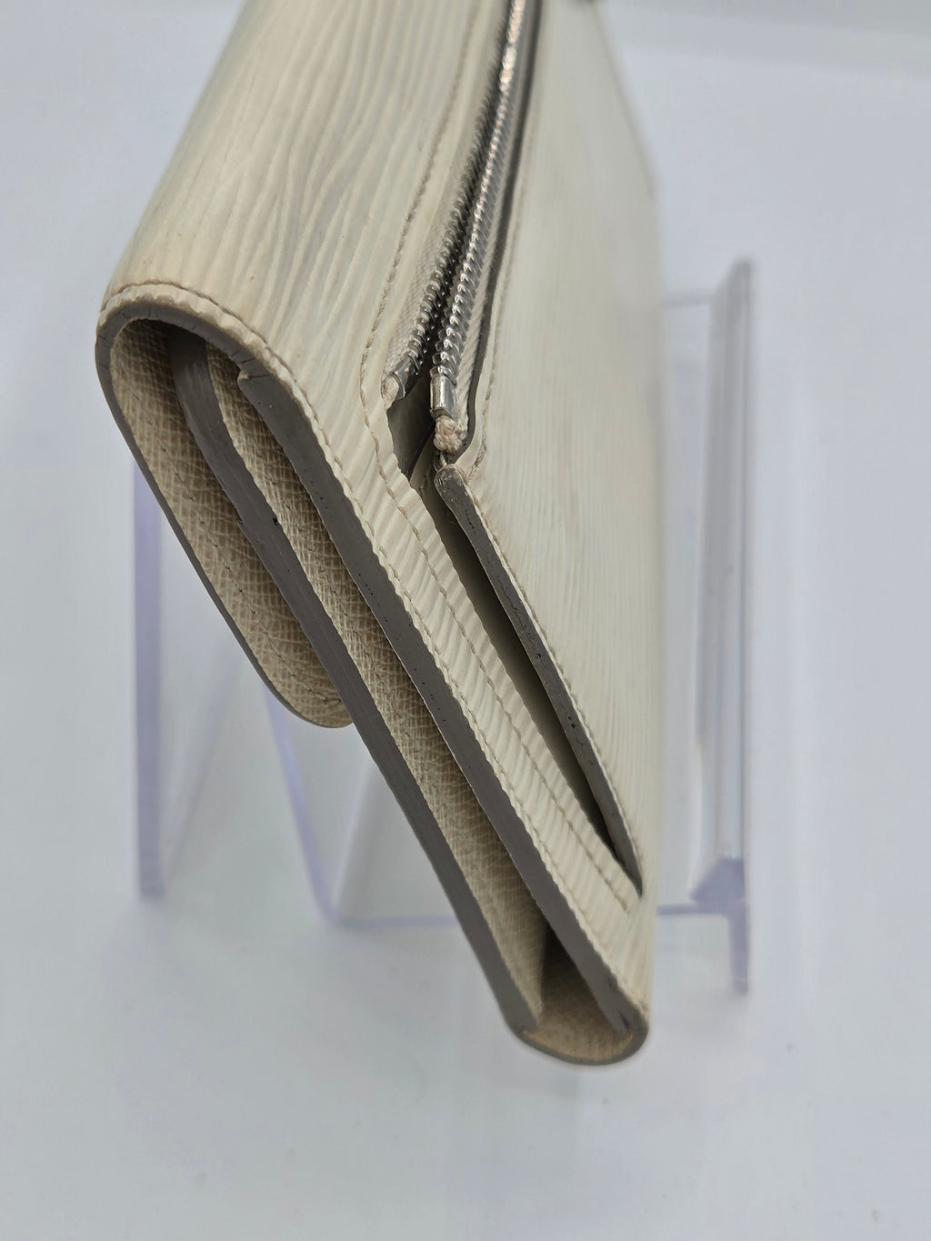 A beige textured leather wallet with a silver zipper, shown folded on a clear acrylic display stand against a white background.