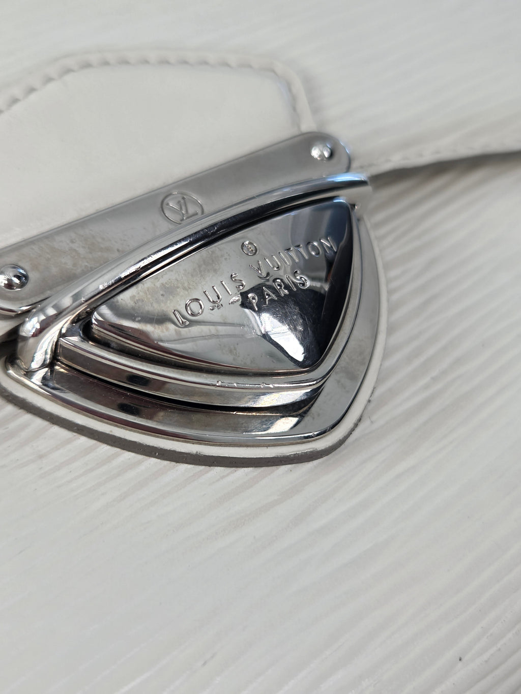 Close-up of a polished silver metal Louis Vuitton monogrammed clasp on a textured white leather surface.