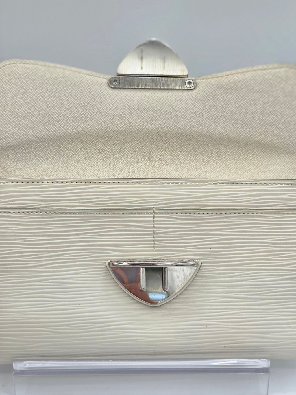 A close-up of a structured handbag in textured cream-colored leather with silver-tone metal hardware, including a top clasp and a central lock.
