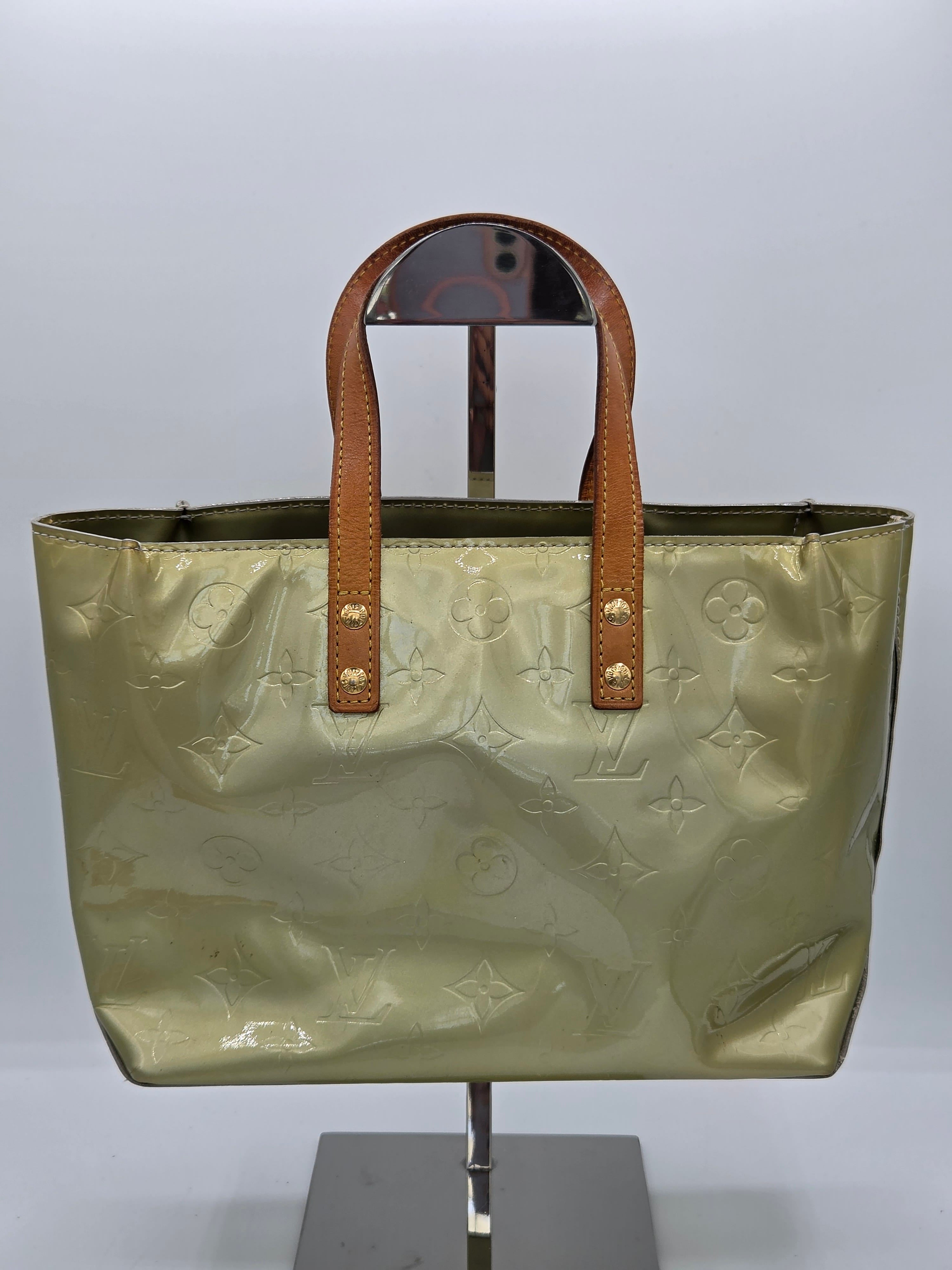 A Louis Vuitton metallic gold monogram tote bag with brown leather handles, displayed on a stand against a light gray background.