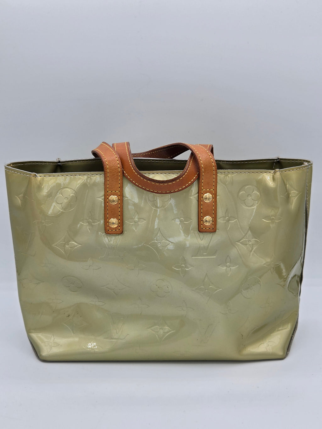 A Louis Vuitton monogram coated canvas tote bag in a metallic olive-gold color, featuring tan leather handles with gold-tone rivets, set against a plain light gray background.