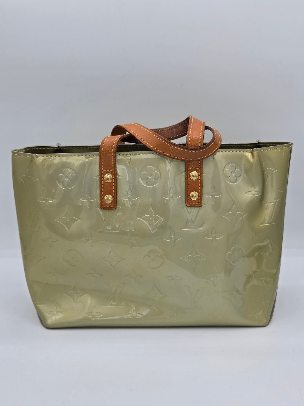 A Louis Vuitton tote bag in metallic olive green patent leather with an embossed monogram pattern, featuring brown leather handles with gold-tone rivets, set against a neutral gray background.