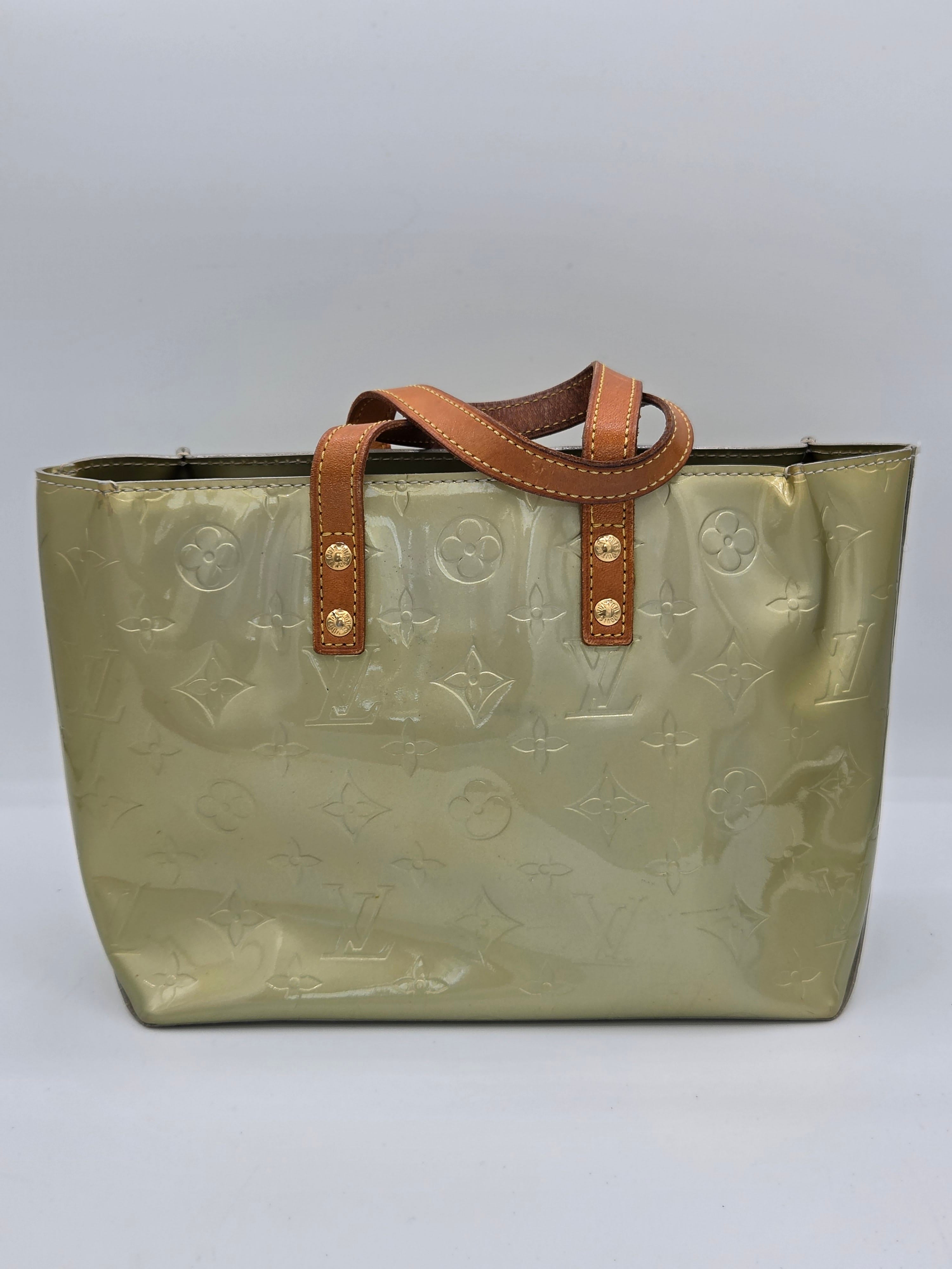 A Louis Vuitton tote bag in metallic olive green patent leather with an embossed monogram pattern, featuring brown leather handles with gold-tone rivets, set against a neutral gray background.