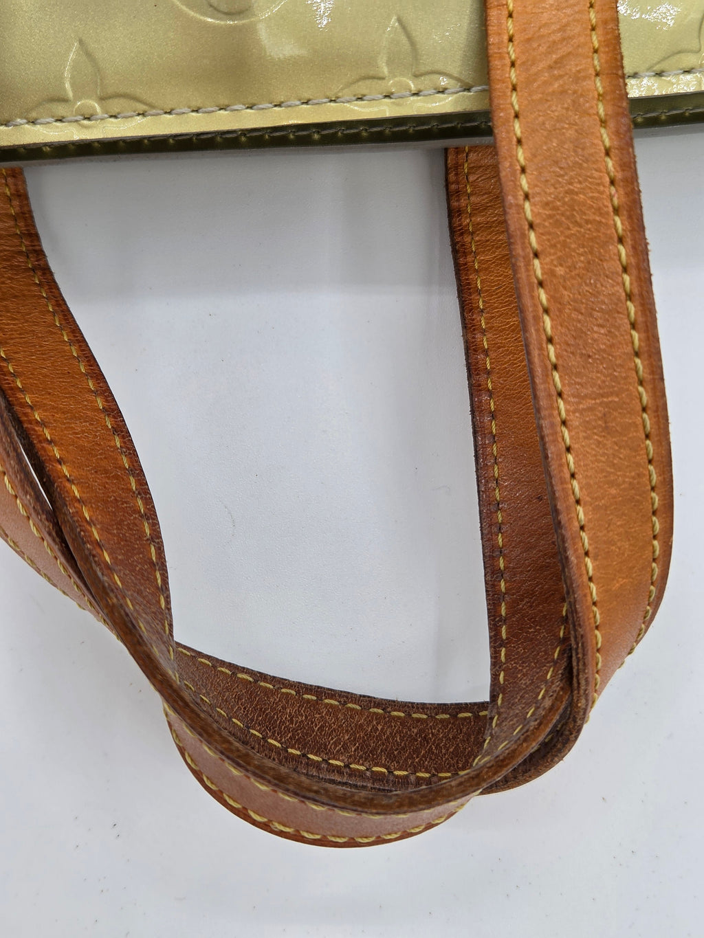 Close-up of a tan leather shoulder strap with light tan contrast stitching, attached to a light beige textured bag body, shown against a plain white background.