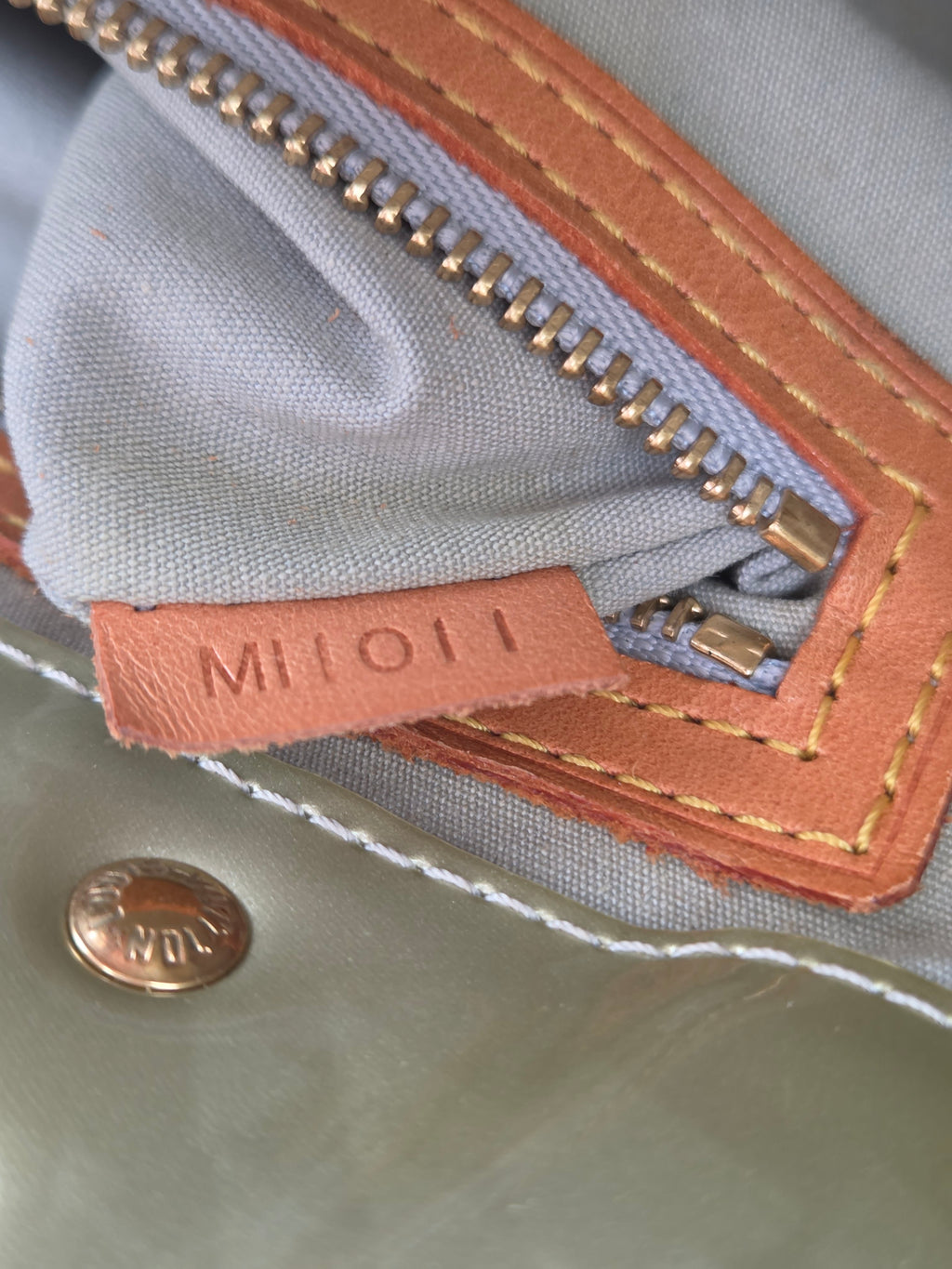 Close-up of a light gray fabric bag with a tan leather zipper pull tab embossed with 'MILIO' and a gold-tone zipper.
