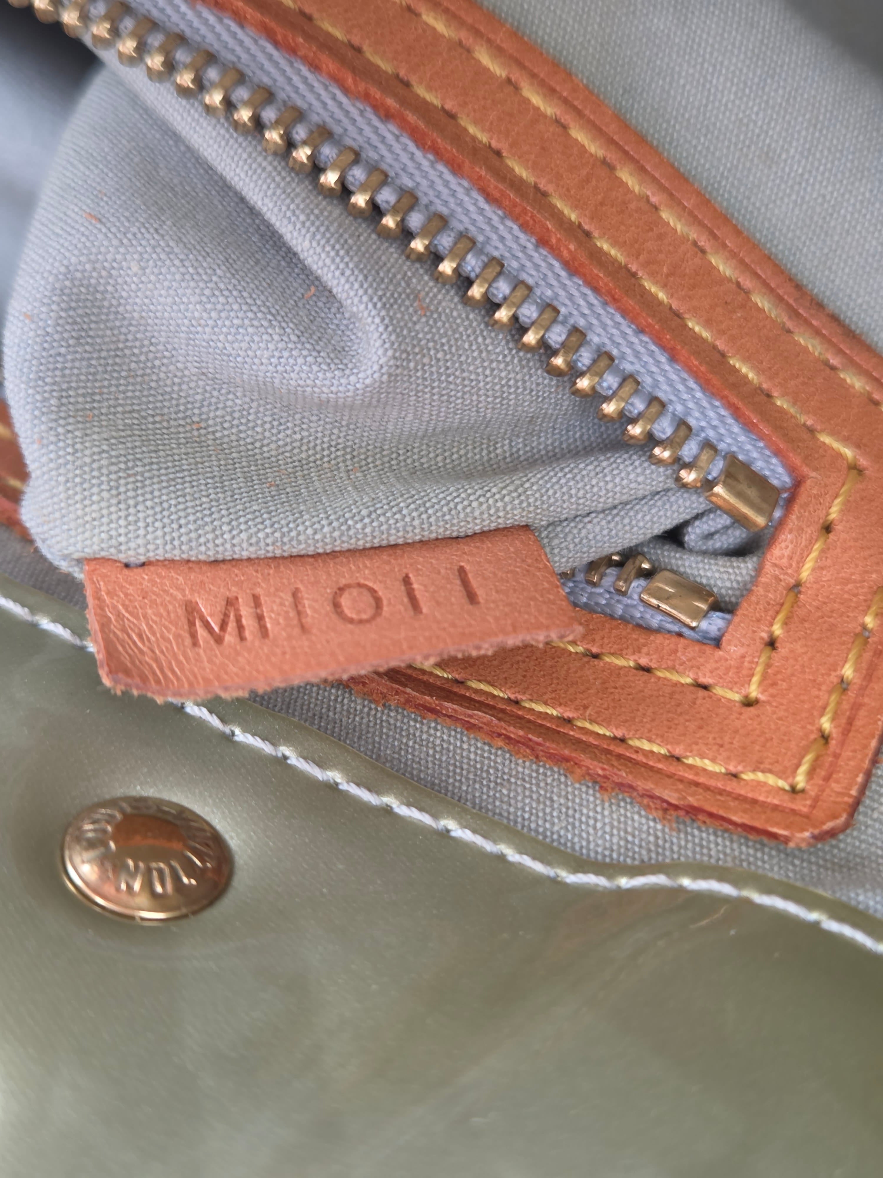 Close-up of a light gray fabric bag with a tan leather zipper pull tab embossed with 'MILIO' and a gold-tone zipper.