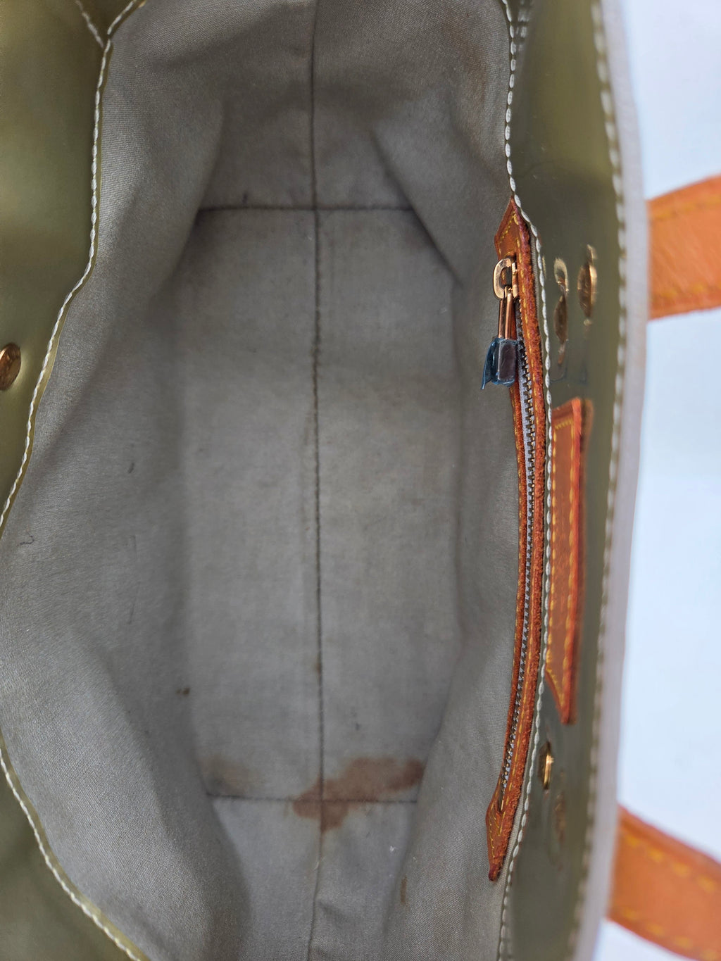 Close-up of the interior of a tote bag, showing a light gray fabric lining, a zipper pocket with tan leather trim, and part of the olive green exterior fabric.