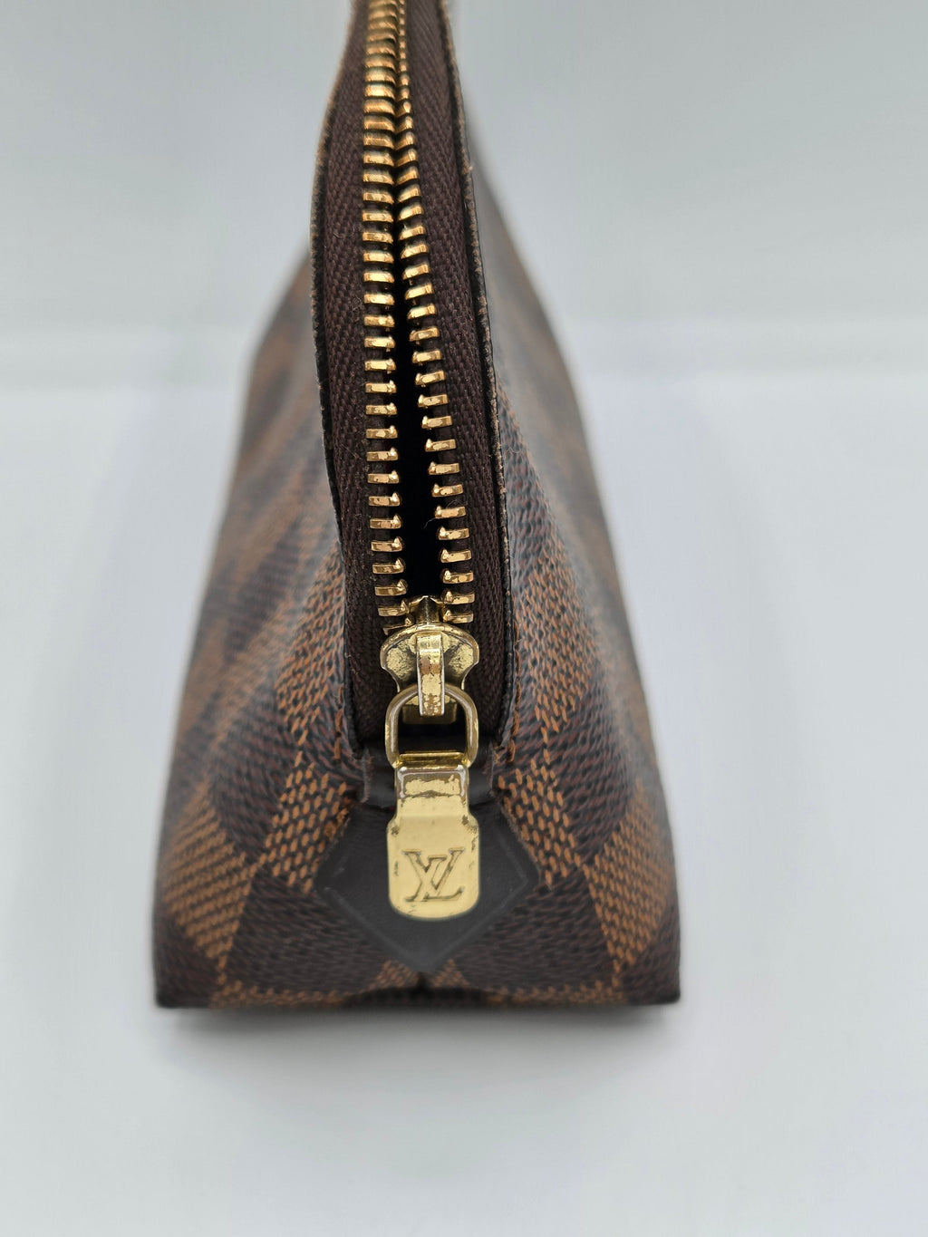 A close-up of a Louis Vuitton pouch in the brand's classic Damier Ebene canvas, featuring a brown and dark brown checkered pattern with a gold-tone zipper and a gold-tone LV logo pull tab.