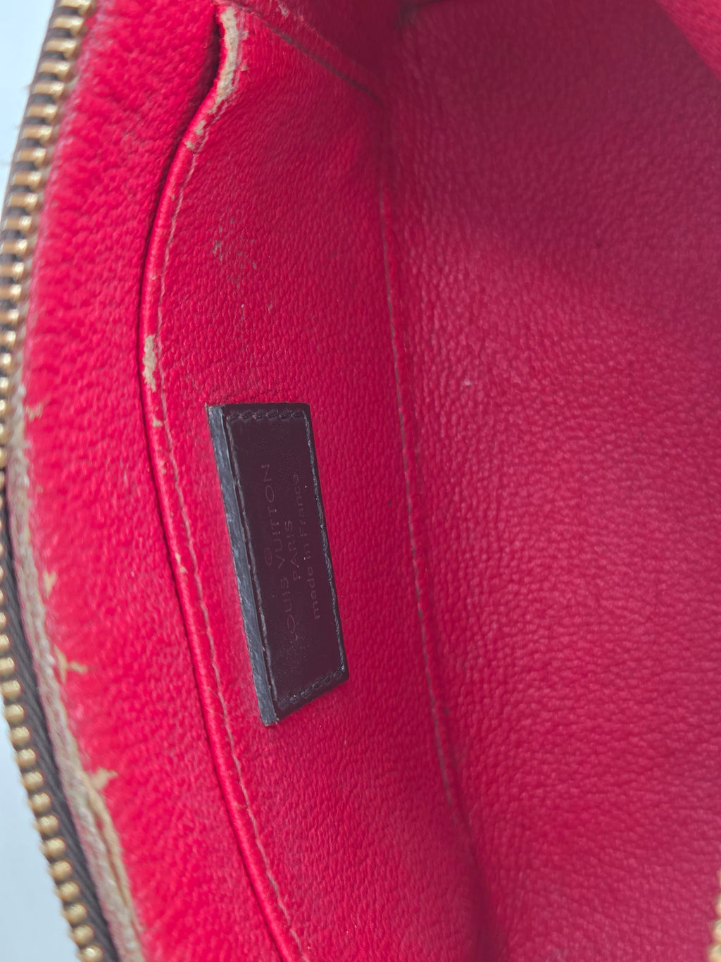 Close-up of the interior lining of a red pebbled leather bag, showing a dark brown rectangular brand patch with embossed text and a gold-tone zipper along the bottom edge.