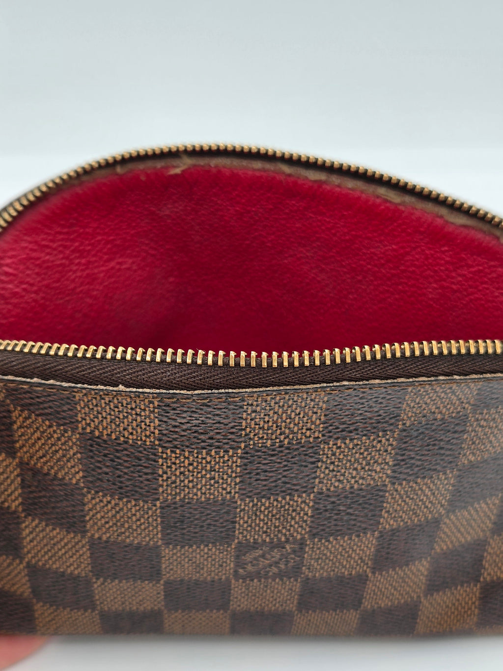 Close-up of a Louis Vuitton Damier canvas pouch showing the brown and beige checkered pattern, a gold zipper, and a red interior lining.