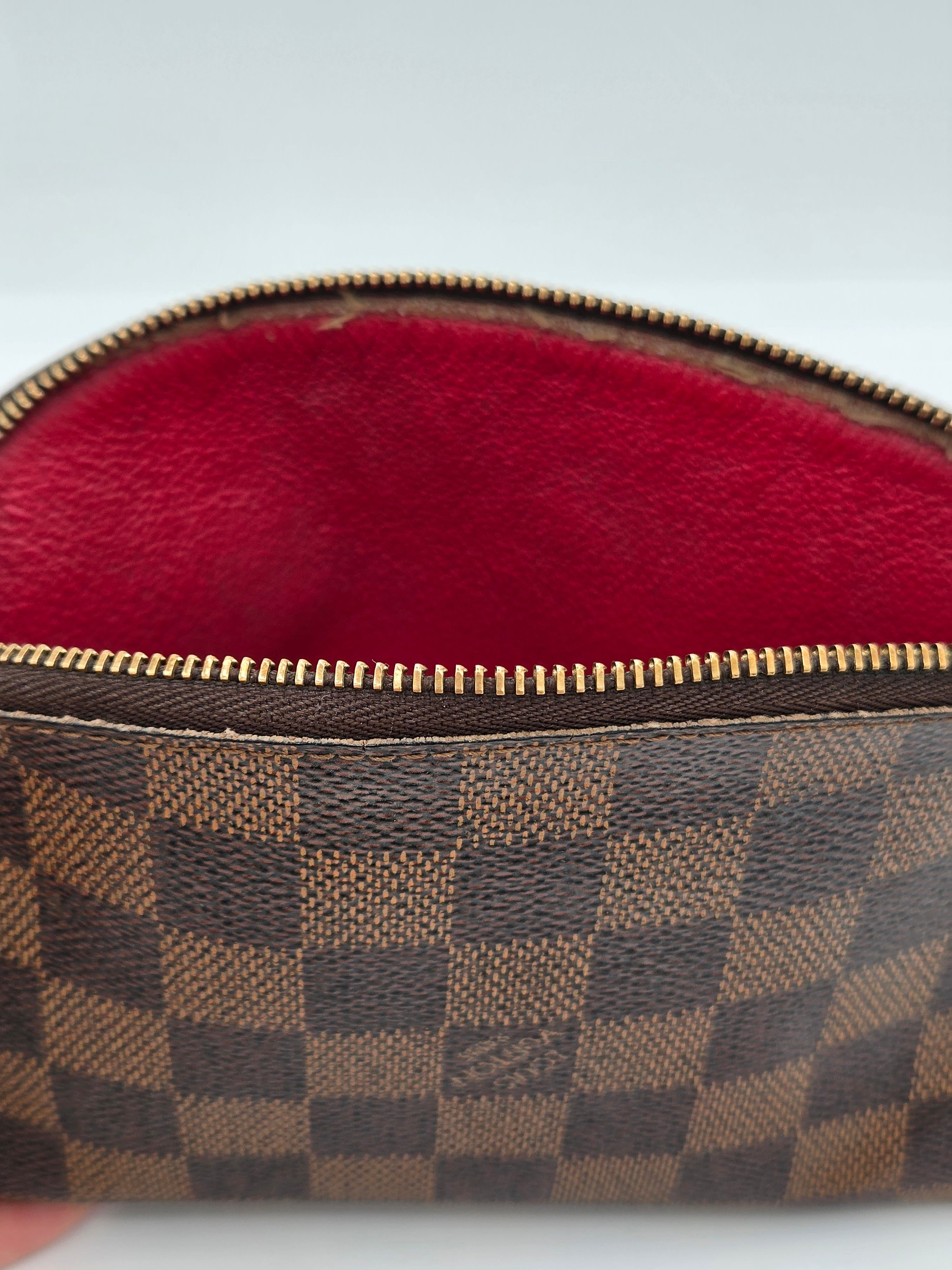 Close-up of a Louis Vuitton Damier canvas pouch showing the brown and beige checkered pattern, a gold zipper, and a red interior lining.