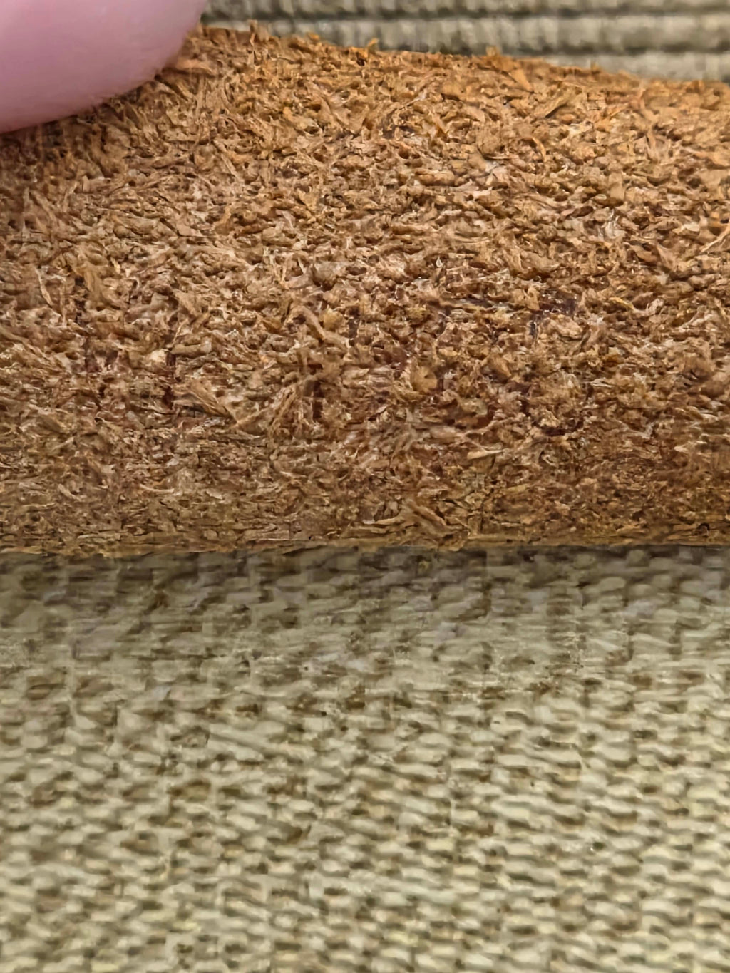 A close-up of a textured brown material, likely a leather or suede roll, held by a finger against a woven beige background.