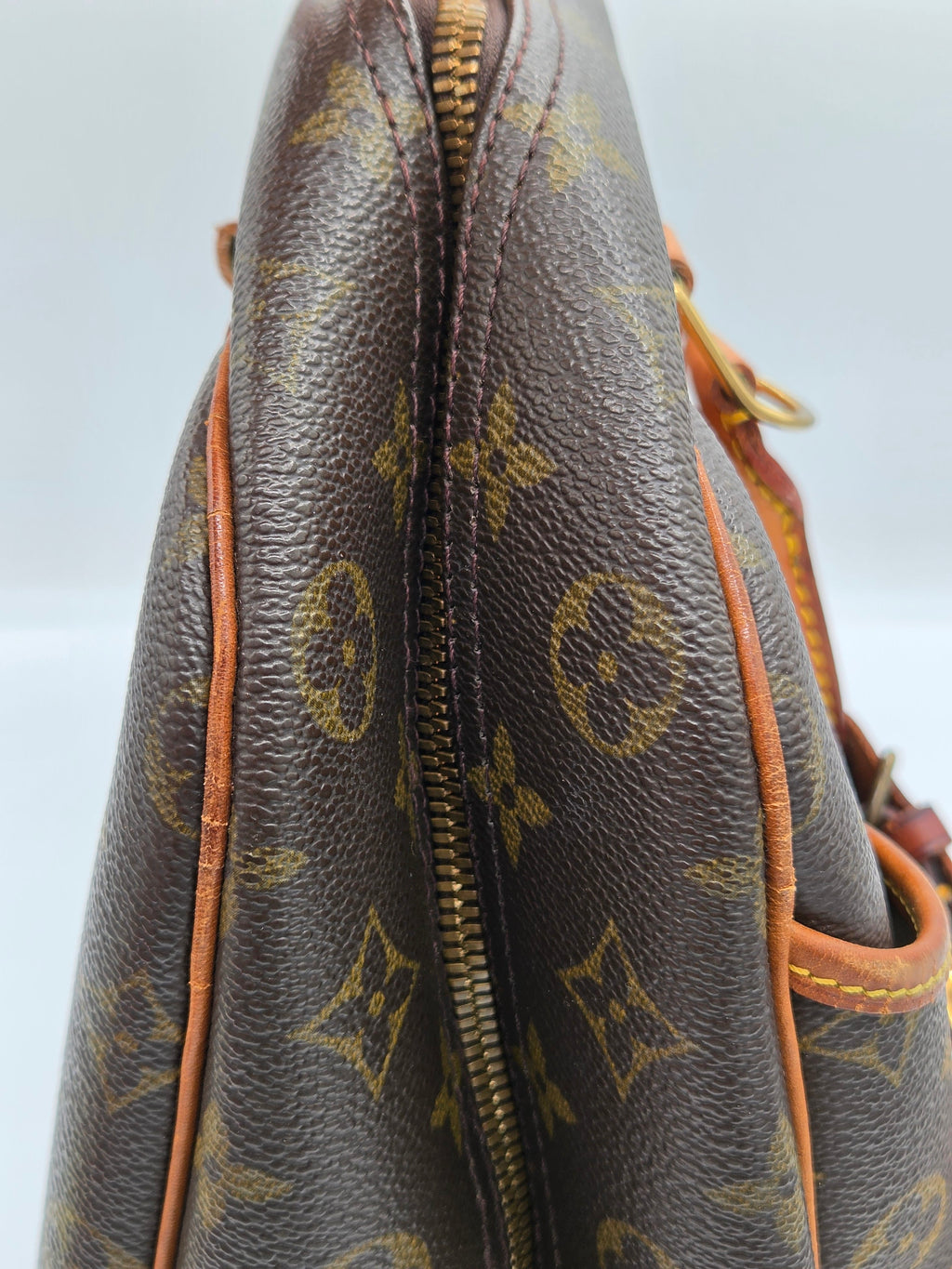 Close-up of a brown monogram canvas handbag with gold-tone hardware and tan leather trim, showing part of the zipper and strap.