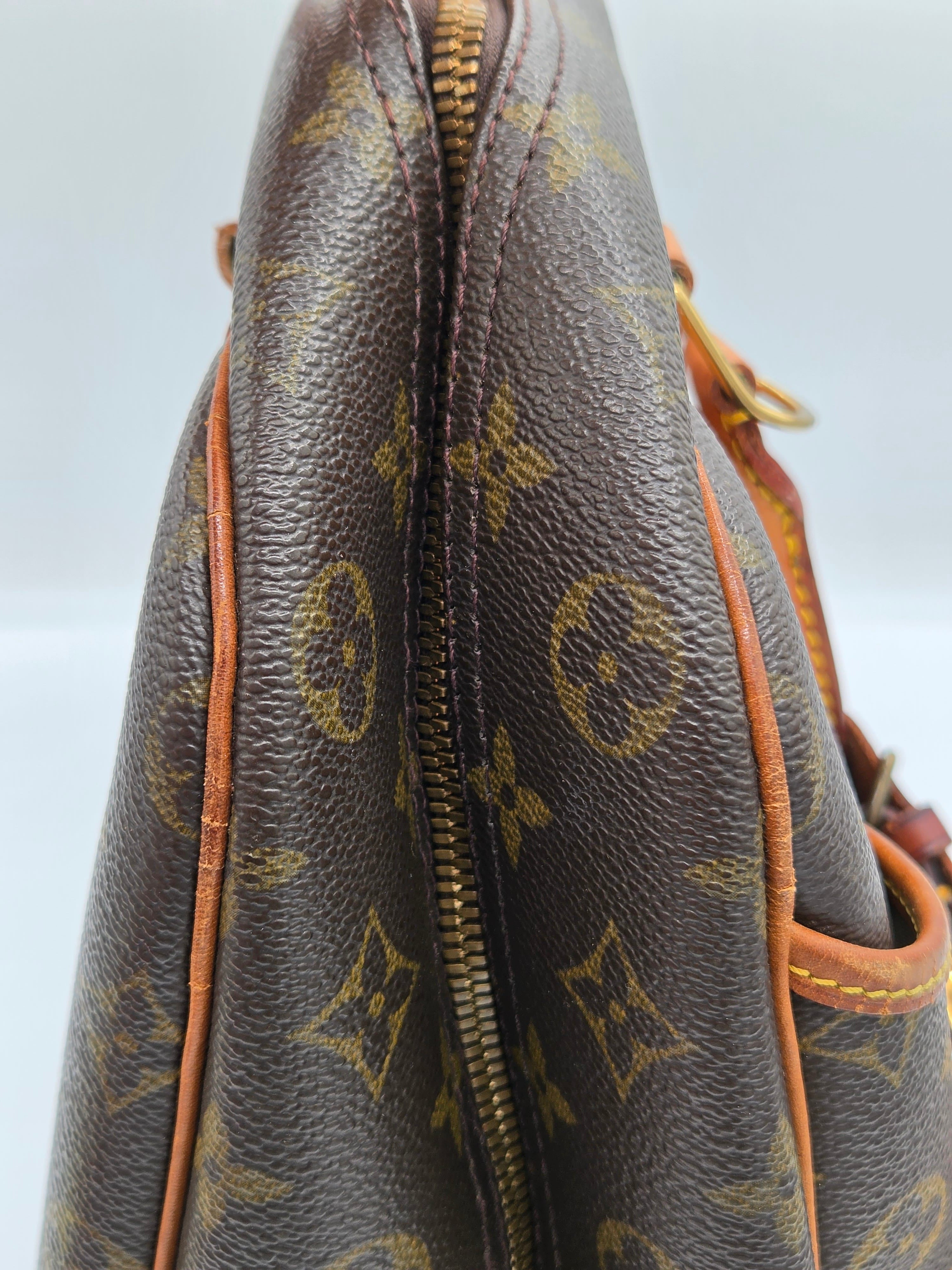 Close-up of a brown monogram canvas handbag with gold-tone hardware and tan leather trim, showing part of the zipper and strap.