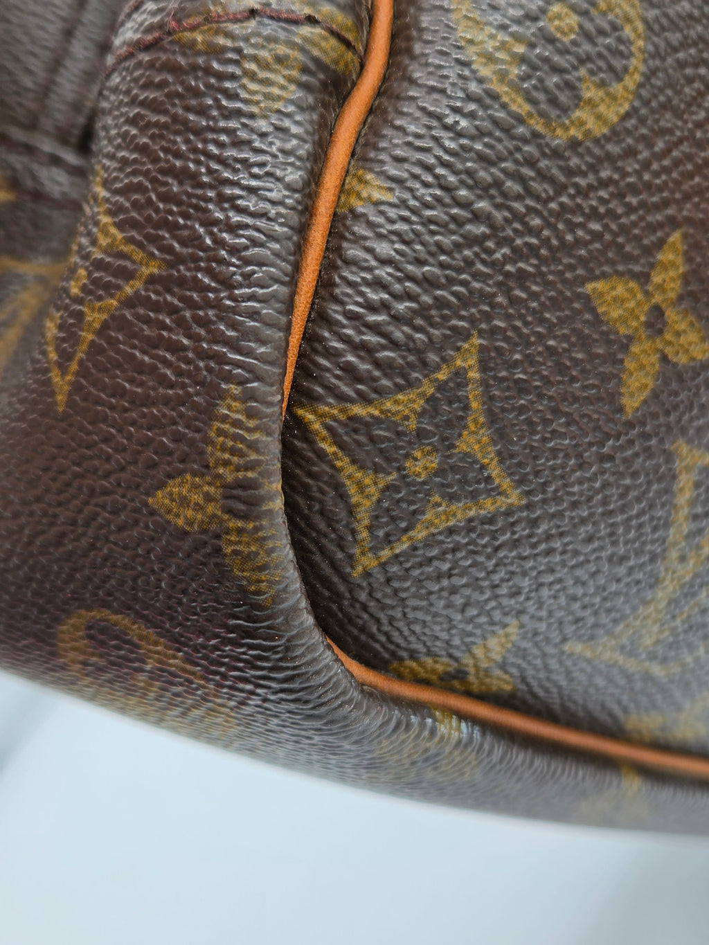 Close-up of a brown coated canvas bag with the classic Louis Vuitton monogram pattern in gold, featuring tan leather piping along the edge.