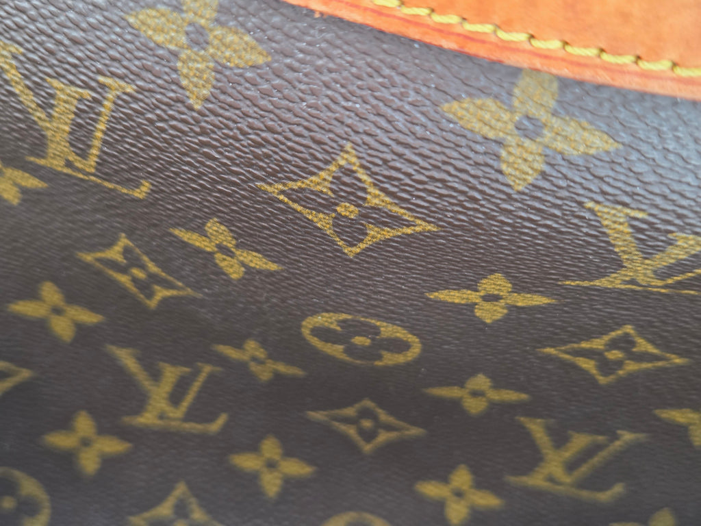 Close-up of Louis Vuitton monogram canvas in dark brown with gold-tone printed monogram motifs and a sliver of tan leather trim at the bottom.