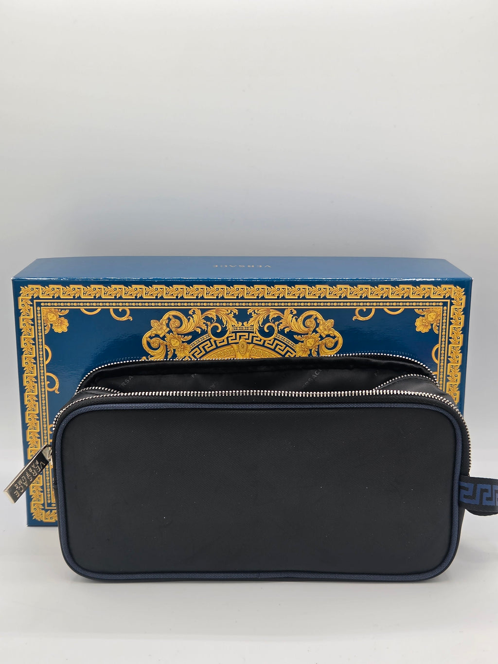 A black zippered cosmetic pouch with a silver zipper, displayed in front of its blue and gold Versace branded box on a light gray background.