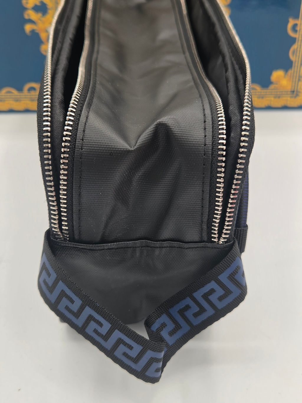 Close-up of a black nylon bag with silver zippers and a blue-gray patterned fabric loop at the bottom, set against a white surface with a blue and gold background.