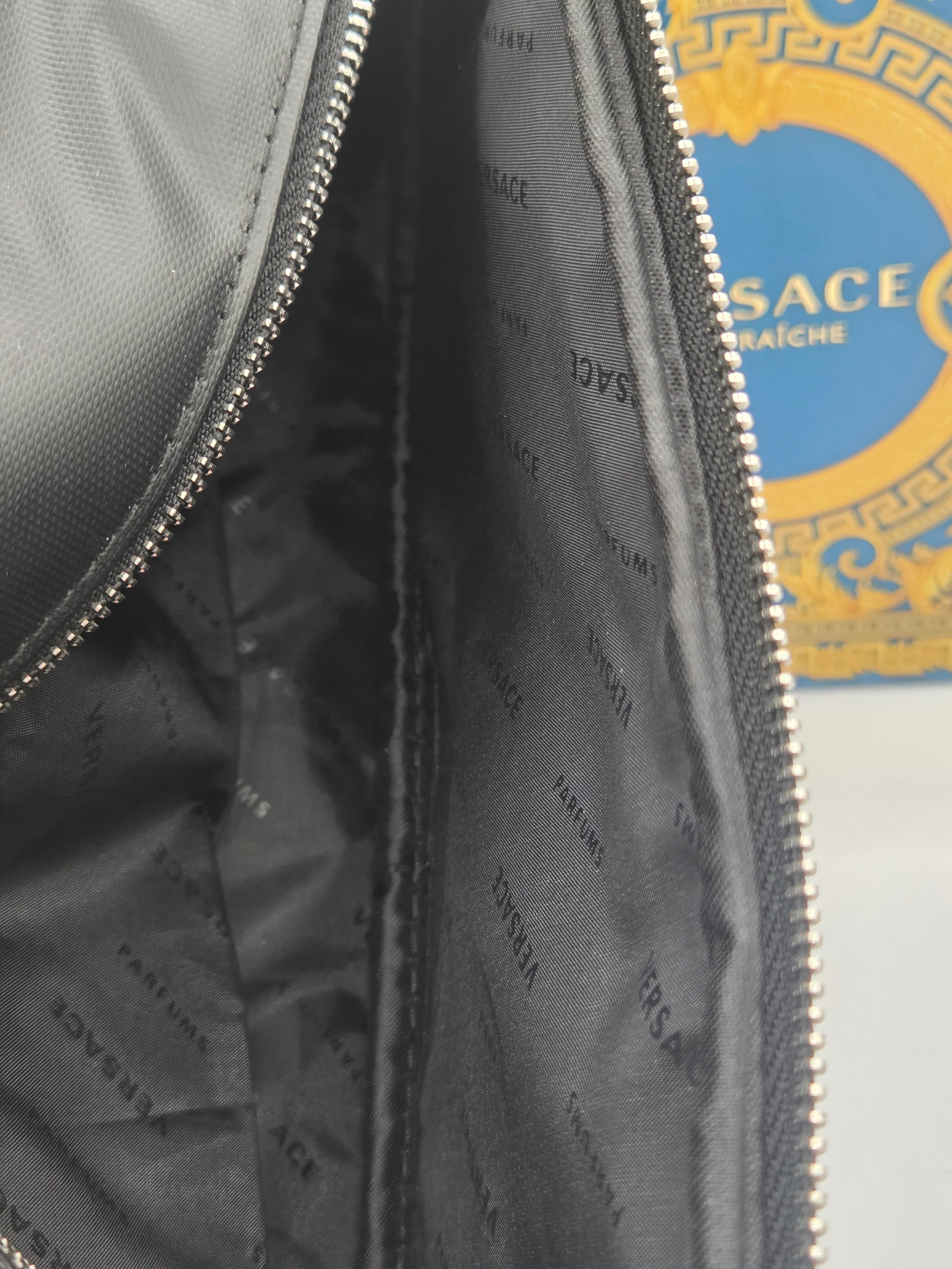 Close-up of a Versace zippered pouch or bag interior, showing the dark gray nylon exterior and branded gray interior lining with silver zipper teeth. A blue and gold Versace logo is partially visible in the background.