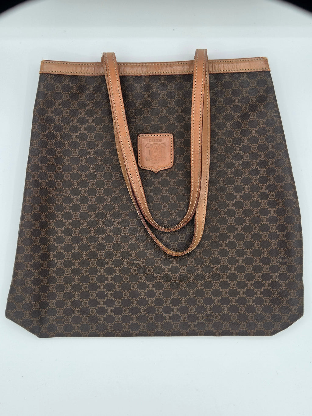 A brown monogram canvas tote bag with tan leather handles and trim, laid flat on a white background.