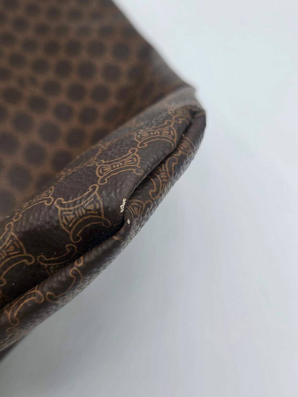 Close-up of a brown patterned fabric item, showing a dark brown surface with a repeating light beige ornamental pattern and a folded edge against a plain light background.