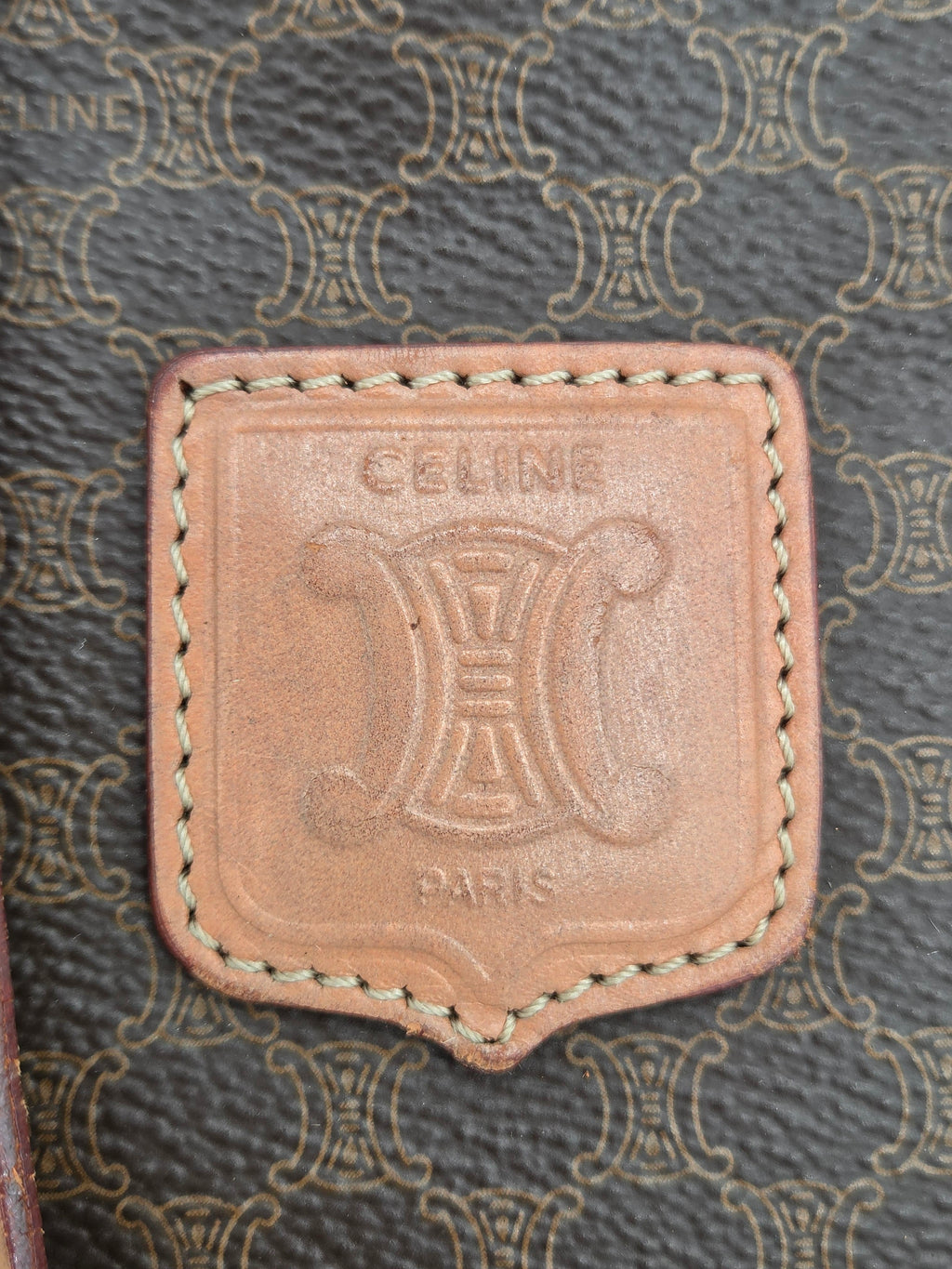Close-up of a Celine leather patch with the brand's signature Triomphe logo and 'CELINE PARIS' text, set against a dark gray patterned background.