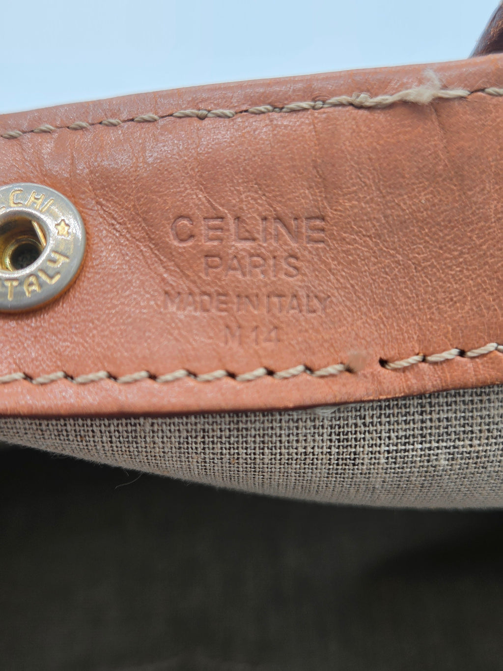 Close-up detail of a Celine shoulder strap featuring tan leather with 'CELINE PARIS MADE IN ITALY' embossed and a silver-tone metal snap button, next to a gray woven fabric.