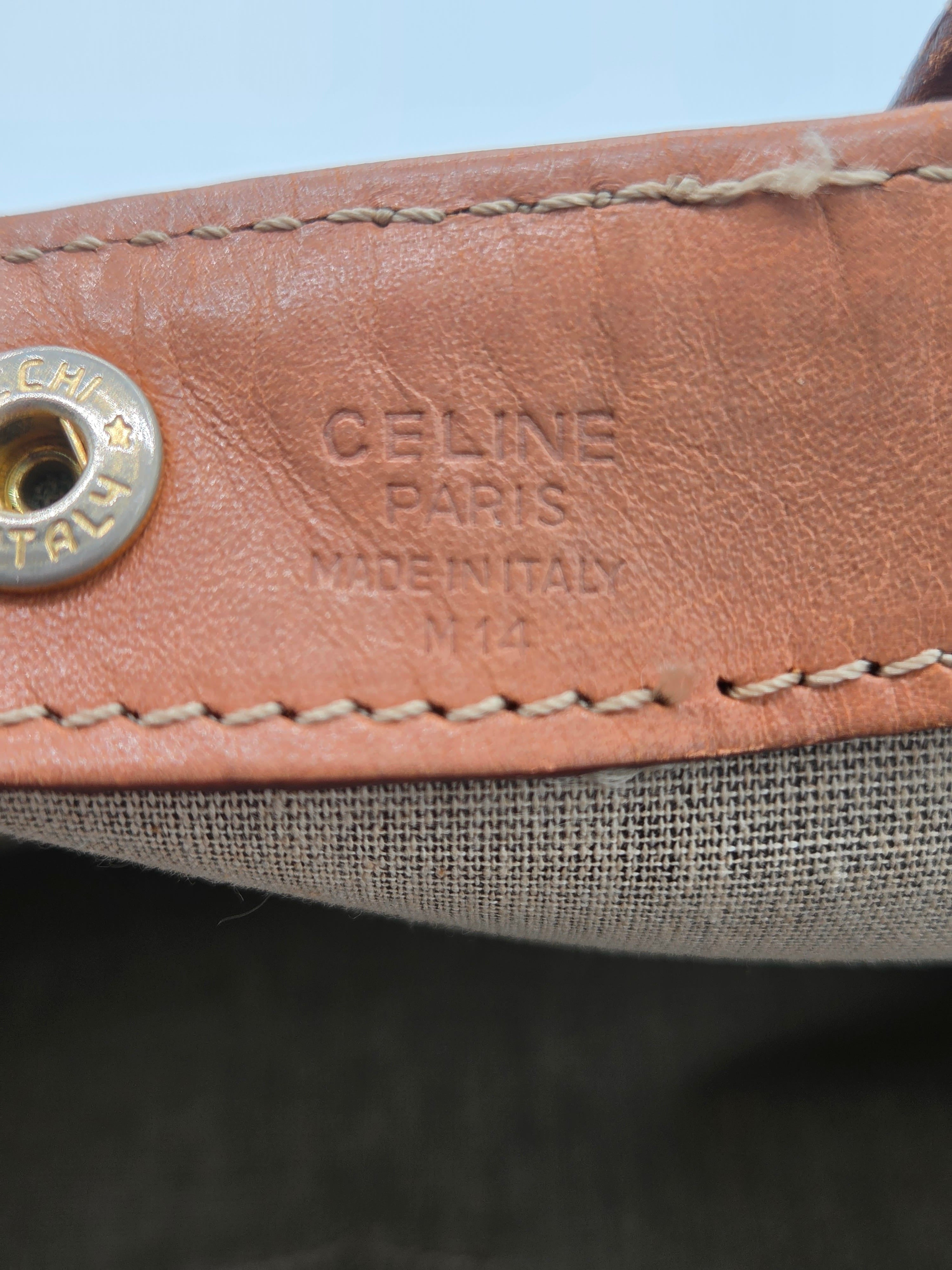 Close-up detail of a Celine shoulder strap featuring tan leather with 'CELINE PARIS MADE IN ITALY' embossed and a silver-tone metal snap button, next to a gray woven fabric.