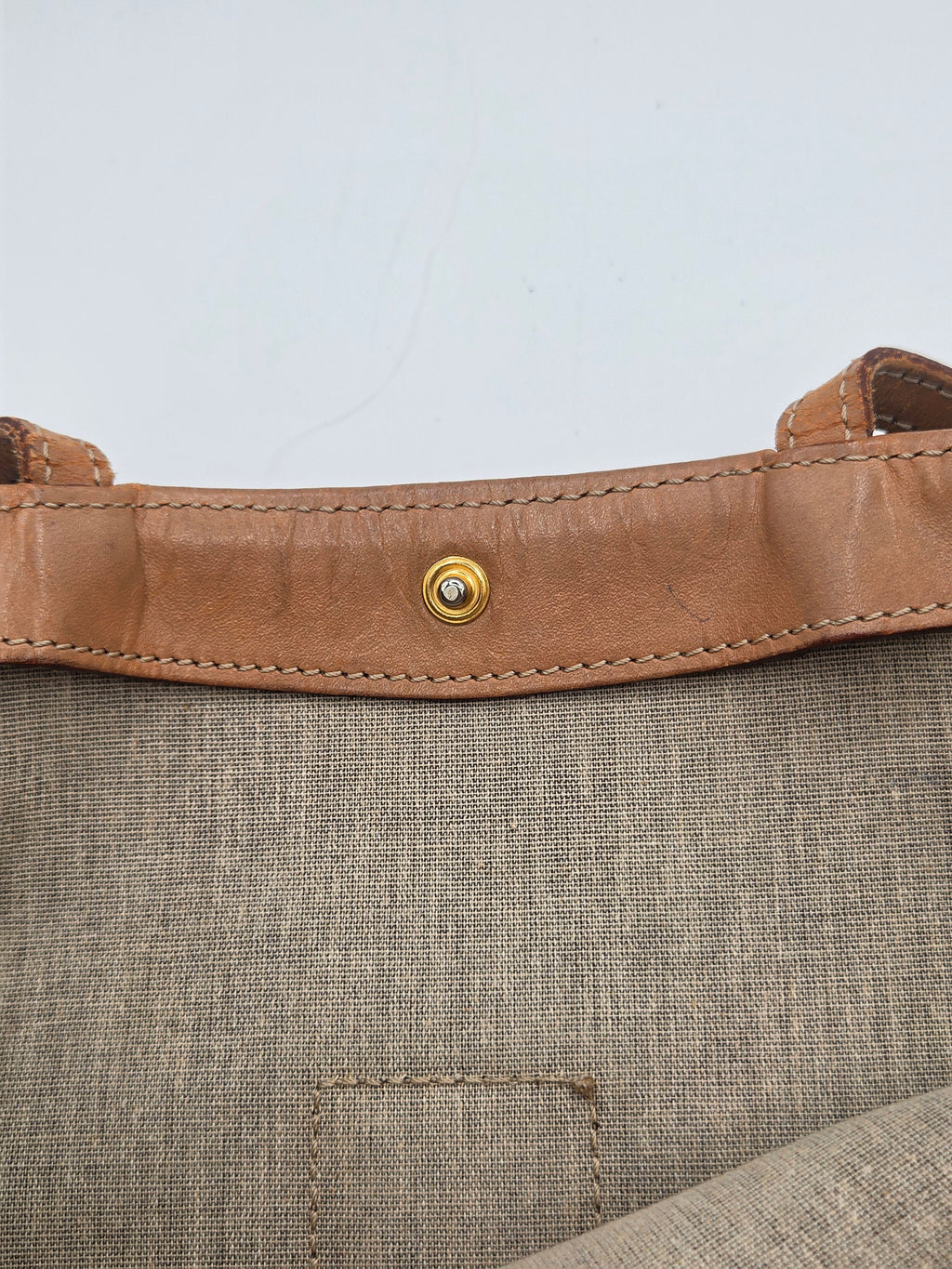Close-up detail of a tan leather strap with a gold-tone snap button, attached to a textured taupe-beige woven fabric panel.