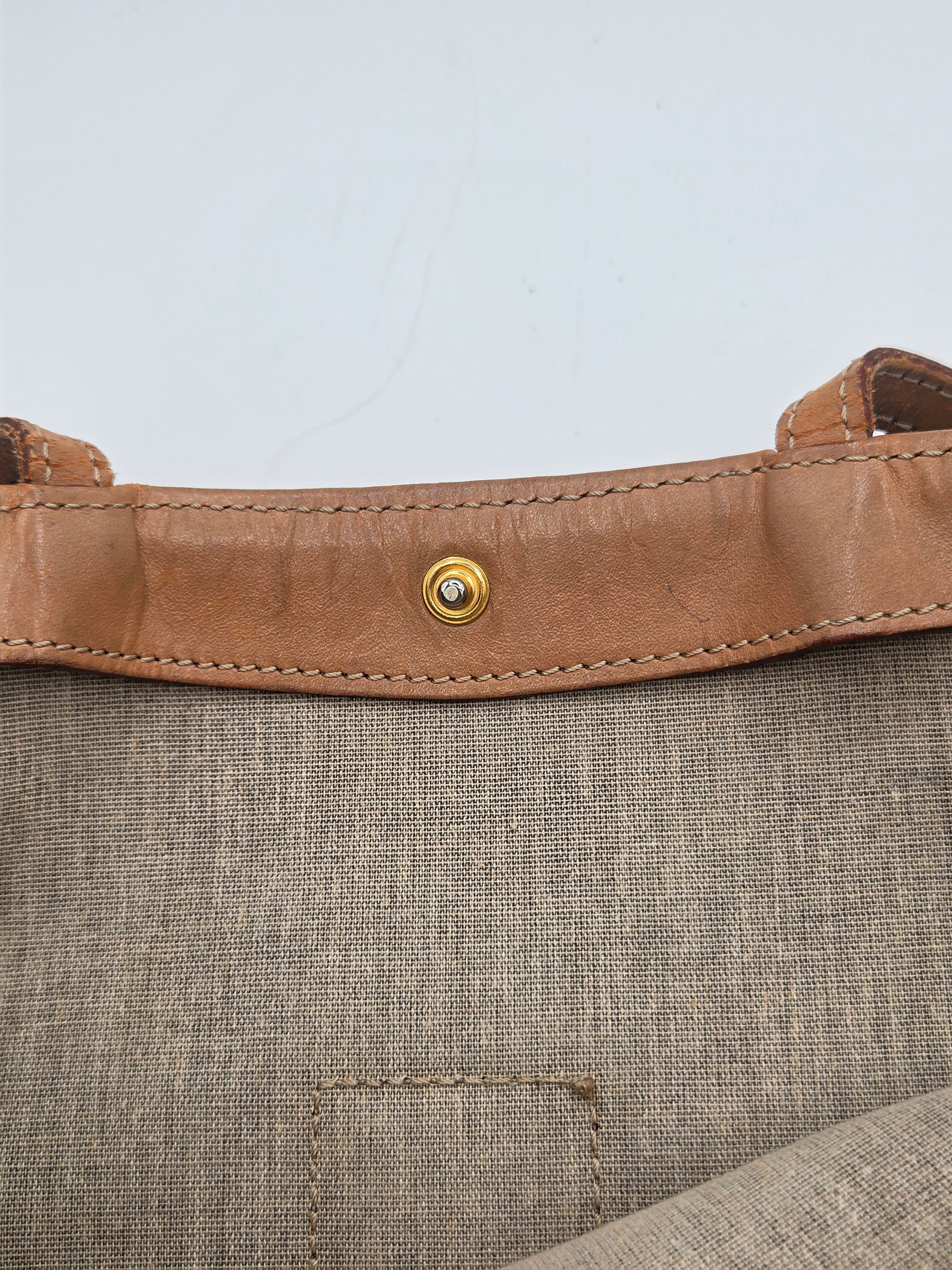 Close-up detail of a tan leather strap with a gold-tone snap button, attached to a textured taupe-beige woven fabric panel.