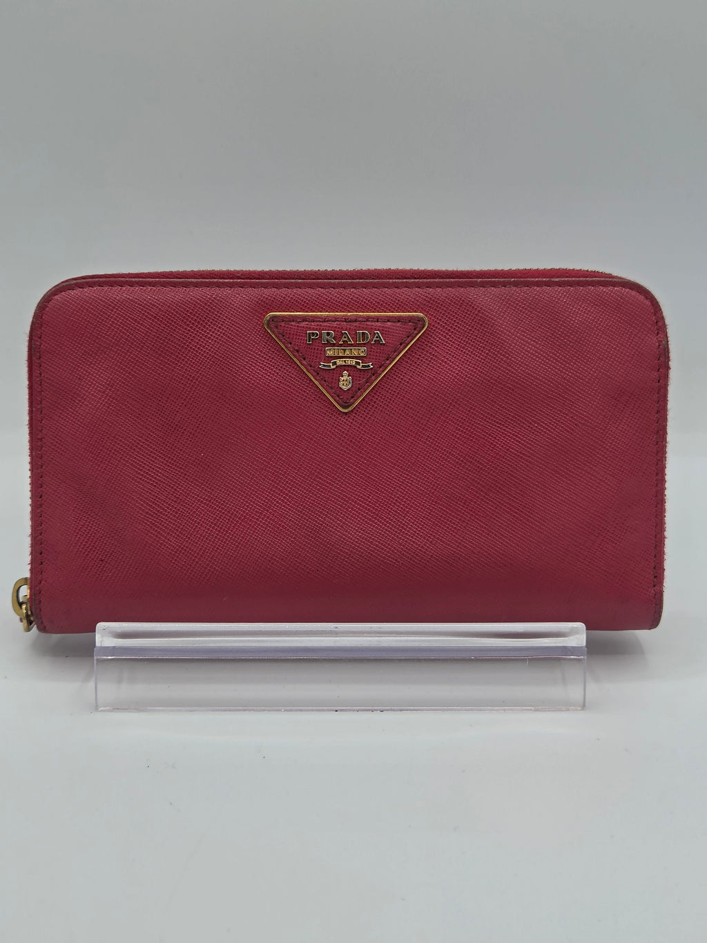 A burgundy Prada zip-around wallet in textured Saffiano leather, featuring the signature gold-tone triangular logo plaque on the front. The wallet is shown standing vertically next to a clear plastic sleeve on a light gray background.