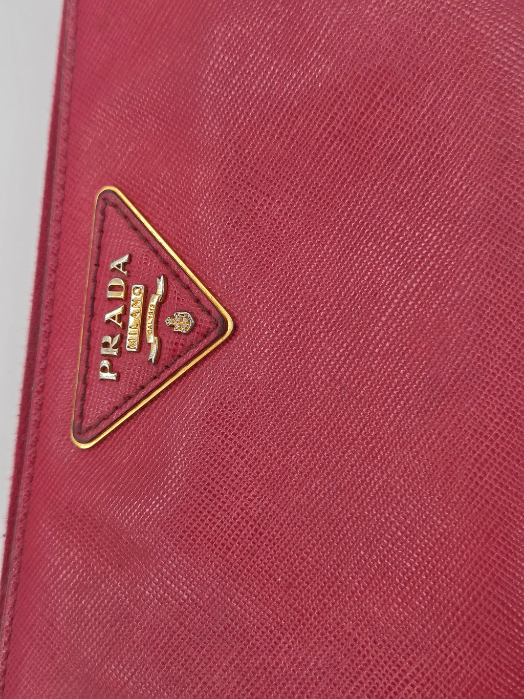 Close-up of a Prada wallet or clutch in textured pinkish-red leather with the signature triangular metal logo plaque on the front.
