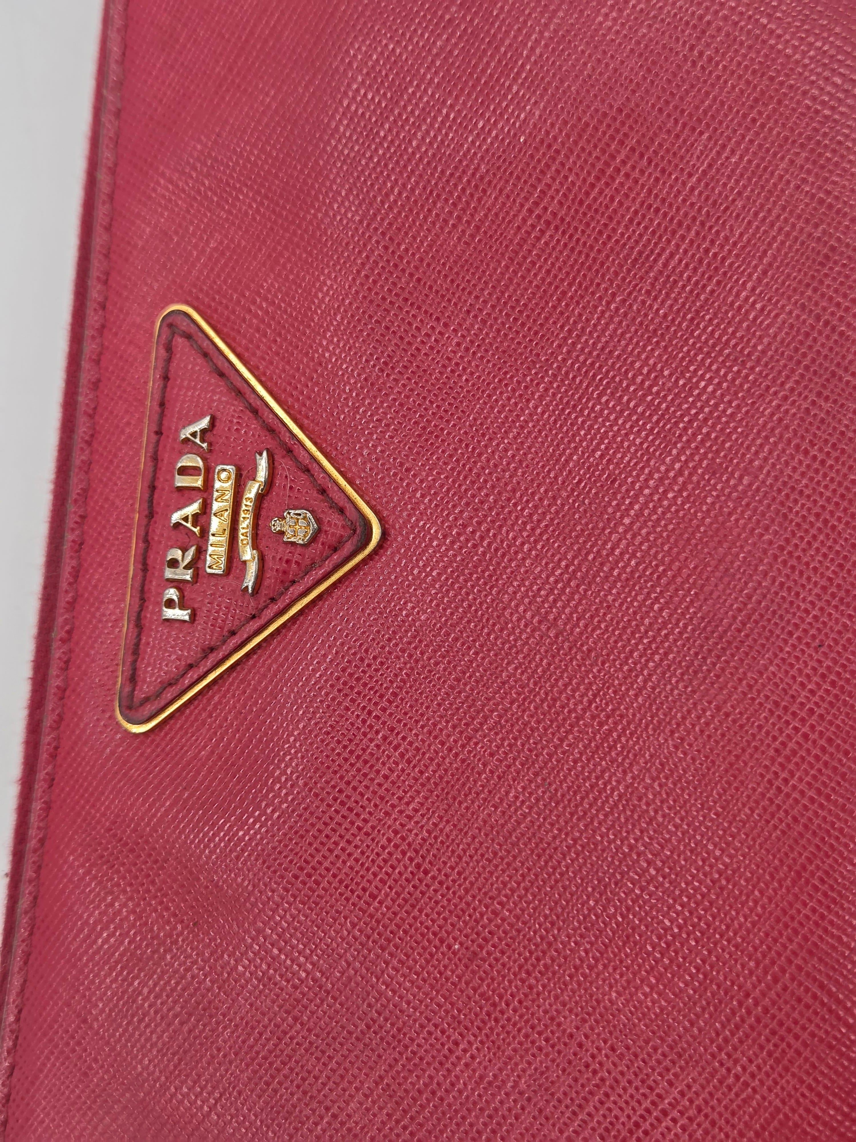 Close-up of a Prada wallet or clutch in textured pinkish-red leather with the signature triangular metal logo plaque on the front.