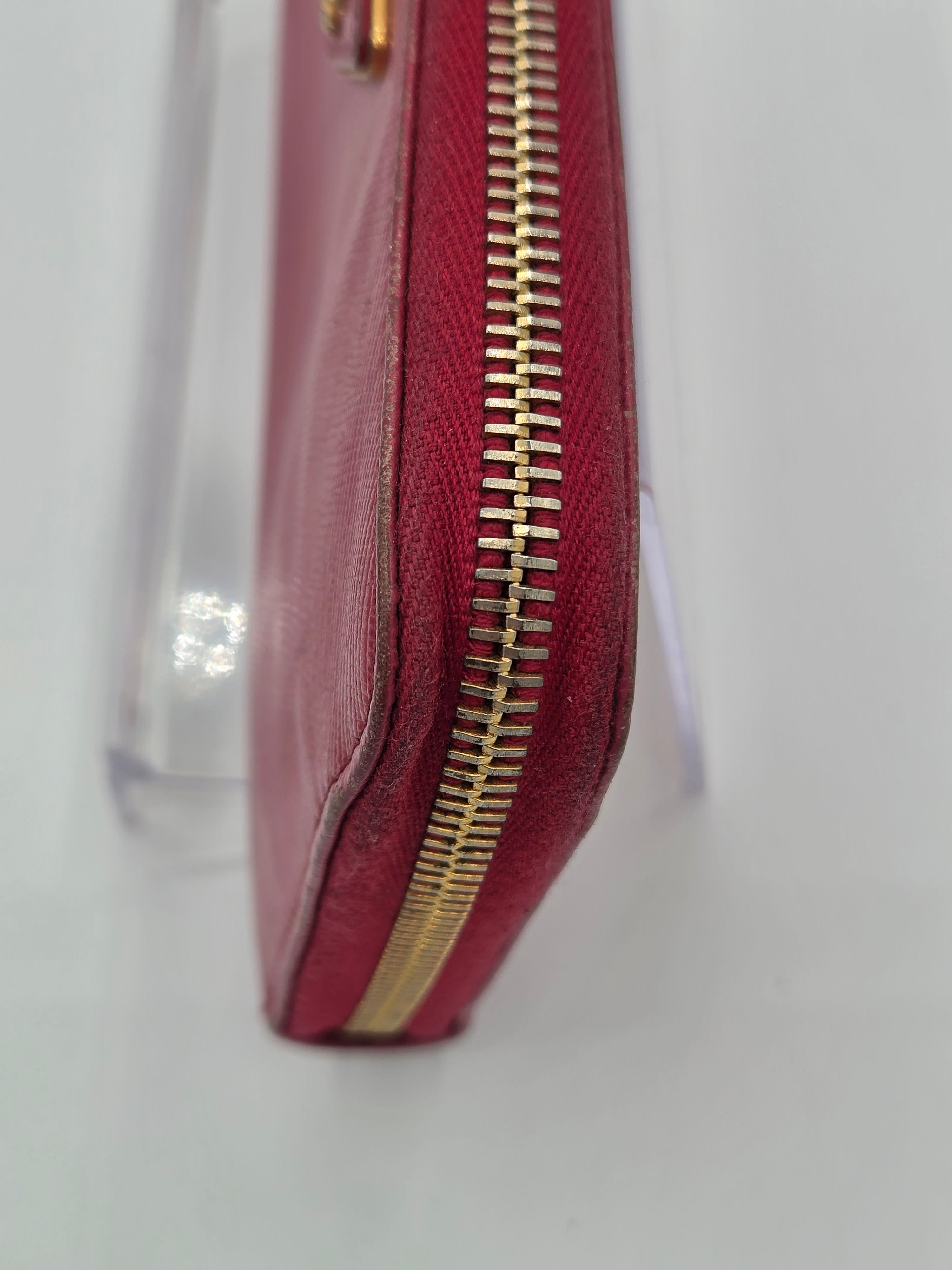 A close-up of a burgundy leather zip-around wallet or clutch with a gold-tone zipper, resting on a clear acrylic stand against a white background.