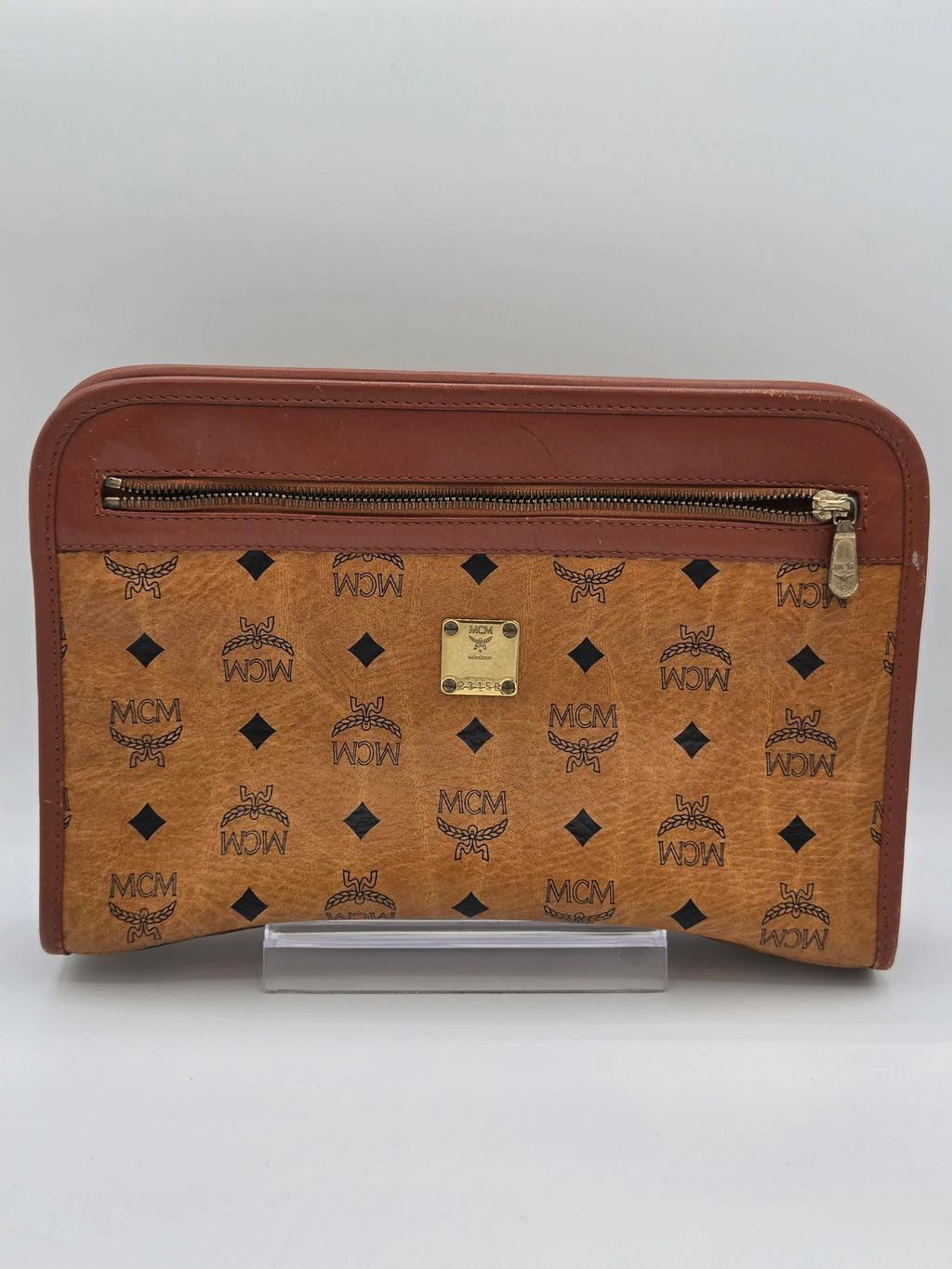 MCM monogram canvas zip-around wallet in tan brown with dark brown leather trim and a gold-tone MCM logo plate, shown closed on a light gray background.